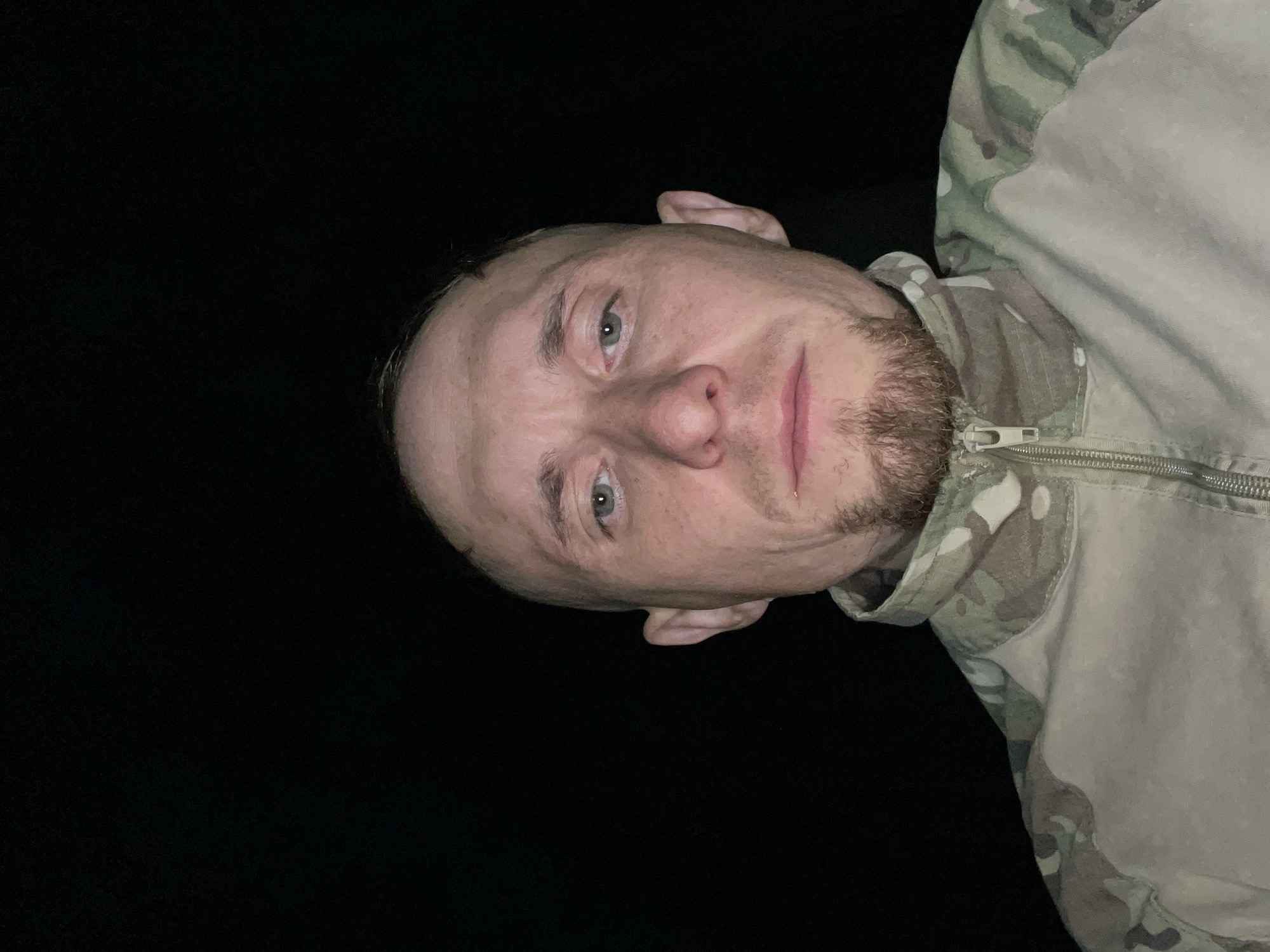 A close-up, front-facing, waist-up selfie of a bald man with a goatee is taken outdoors at night. The man is wearing a camouflage jacket with a zippered collar. His expression is neutral, and he is looking directly at the camera. The background is completely black, indicating the absence of any discernible environmental features. The lighting appears to be coming from the front, illuminating the man's face and upper body. The location is identified as Nikolayevka, Russia.