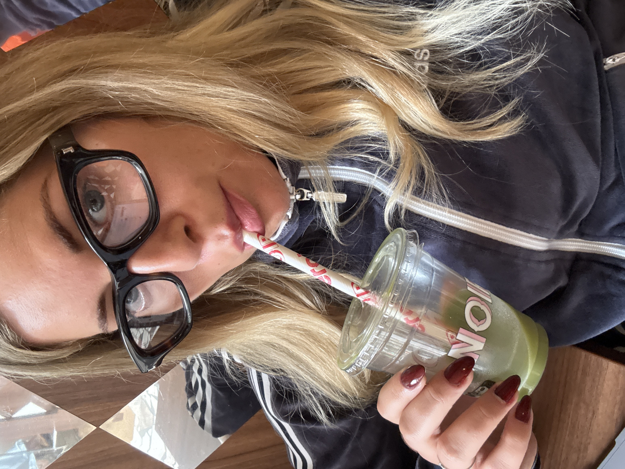 A woman with long blonde hair is holding a clear plastic cup with a green drink and a red and white striped straw. She is wearing black glasses and a dark blue jacket with white stripes on the sleeves. Her nails are painted dark red. The cup has a pink and white logo on it. The surface below is a patterned brown and white, possibly tiled. The image appears to be taken indoors, possibly at a cafe or restaurant. The location in the city of Paris, France cannot be identified based on this image.