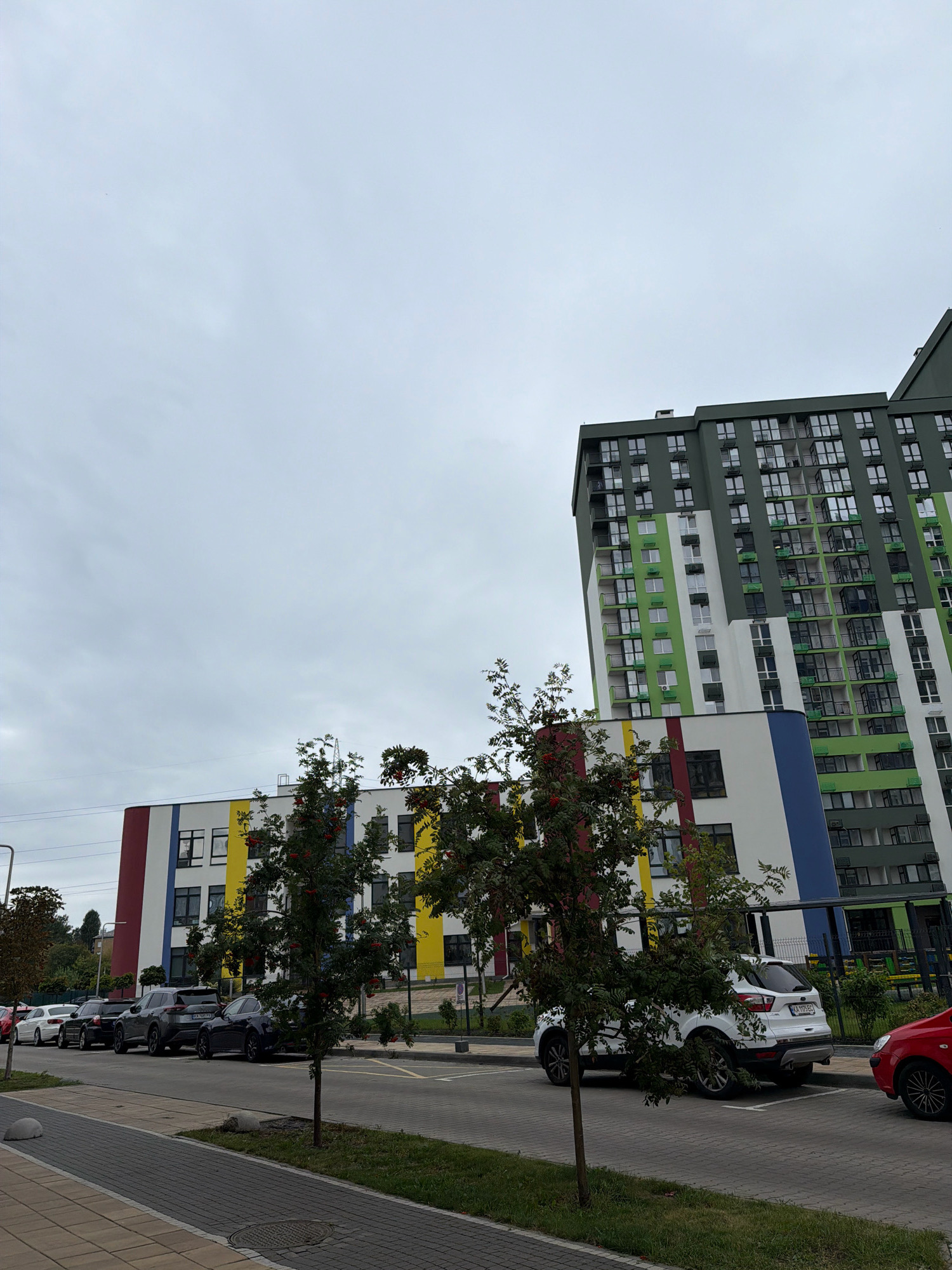 In Kyiv, Ukraine, on a cloudy day, a street scene unfolds with modern residential buildings and parked cars. A multi-story building with a grey and green facade dominates the right side of the frame, featuring numerous windows and balconies. To its left, a lower, more colorful building displays sections of white, yellow, blue, and red. Trees with red berries, likely rowan trees, are positioned in front of these structures. Several cars are parked along the street, including a white SUV, a dark sedan, and other vehicles partially visible. The sidewalk is paved with grey and beige interlocking bricks, and a patch of green grass borders the street. In the background, power lines are visible against the overcast sky.