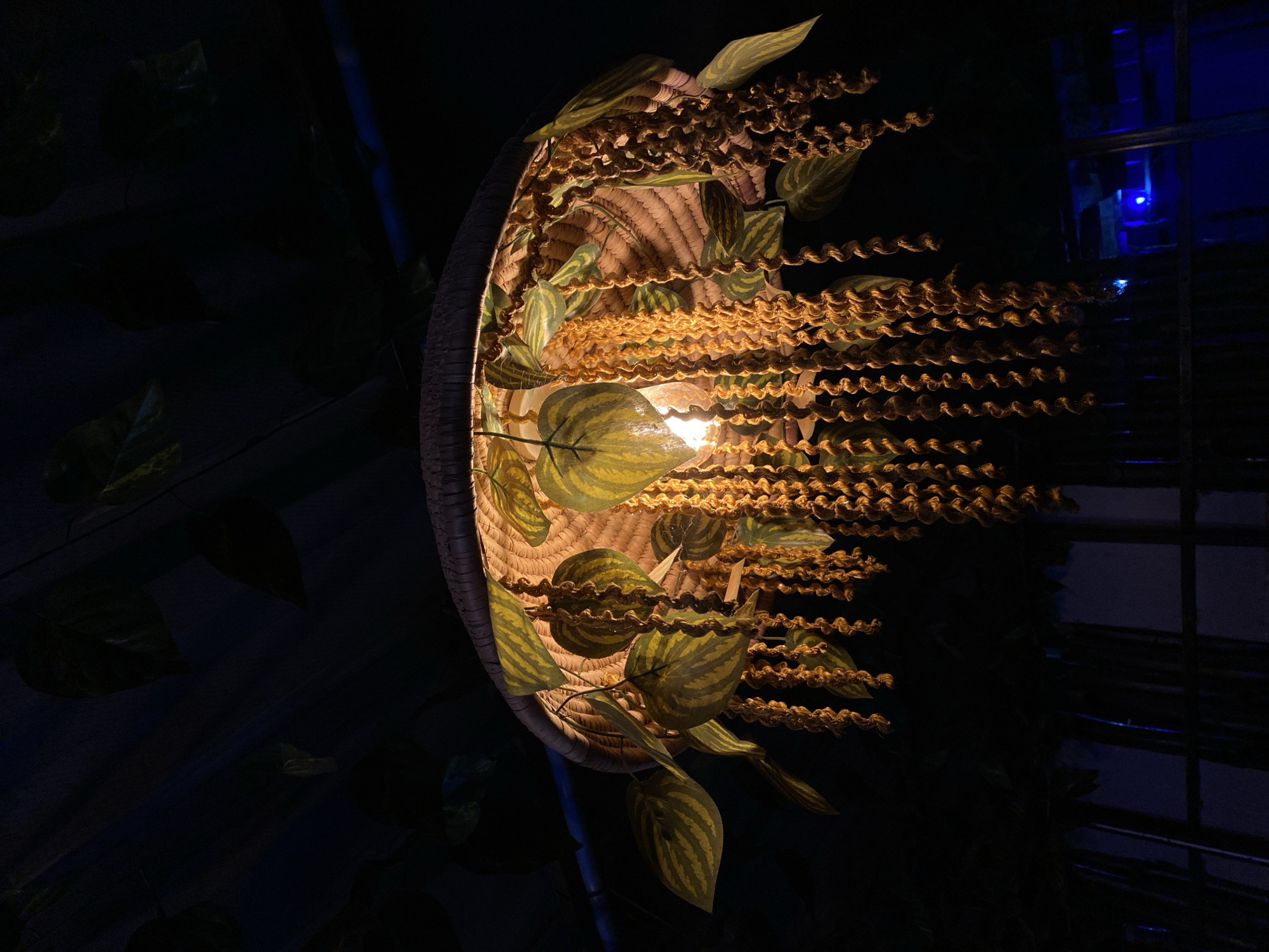 The image shows a decorative hanging lamp with an illuminated bulb. The lamp is made of a woven material, possibly straw, and is adorned with artificial leaves and hanging strands of coiled material. The lamp is suspended from a dark ceiling, possibly wooden planks, with other foliage visible in the background. A blue light is visible in the lower right portion of the image, suggesting another light source or illuminated object in the distance. The overall scene appears to be indoors, likely in a dimly lit environment. No people or specific actions are depicted. The image does not contain any information to infer its location in Gura Top, Nigeria.