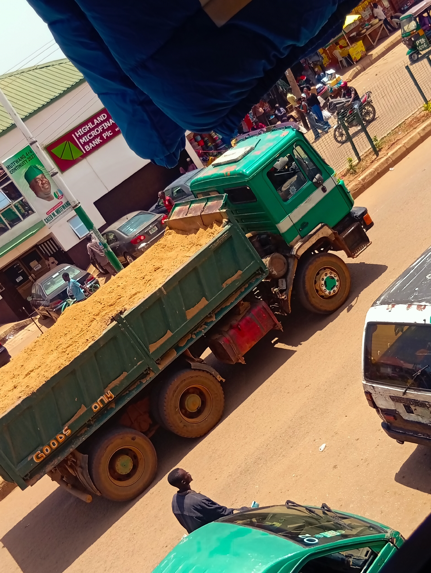 In Bukuru, Nigeria, a bustling scene unfolds on a sun-drenched street. A large green dump truck, its bed filled with light brown sand, is prominently positioned. On its side, "GOODS ONLY" is emblazoned in faded yellow lettering. To the right, the rear of a white van is visible, its paintwork weathered. In the foreground, a man in a dark shirt stands, looking upwards. Behind the dump truck, a dark sedan is parked, and a person in a lighter blue shirt can be seen leaning into its trunk. Further back, a green and white sign with the Highland Microfinance Bank Plc logo is affixed to a building. A promotional poster featuring a man in a green cap and a candidate's name is also visible. The street is lined with shops, some with yellow awnings, and the area appears to be a commercial hub. In the background, a fence separates the street from a bustling market area with people browsing stalls. A motorcycle and a three-wheeled vehicle are also present on the street, suggesting a lively and active urban environment.