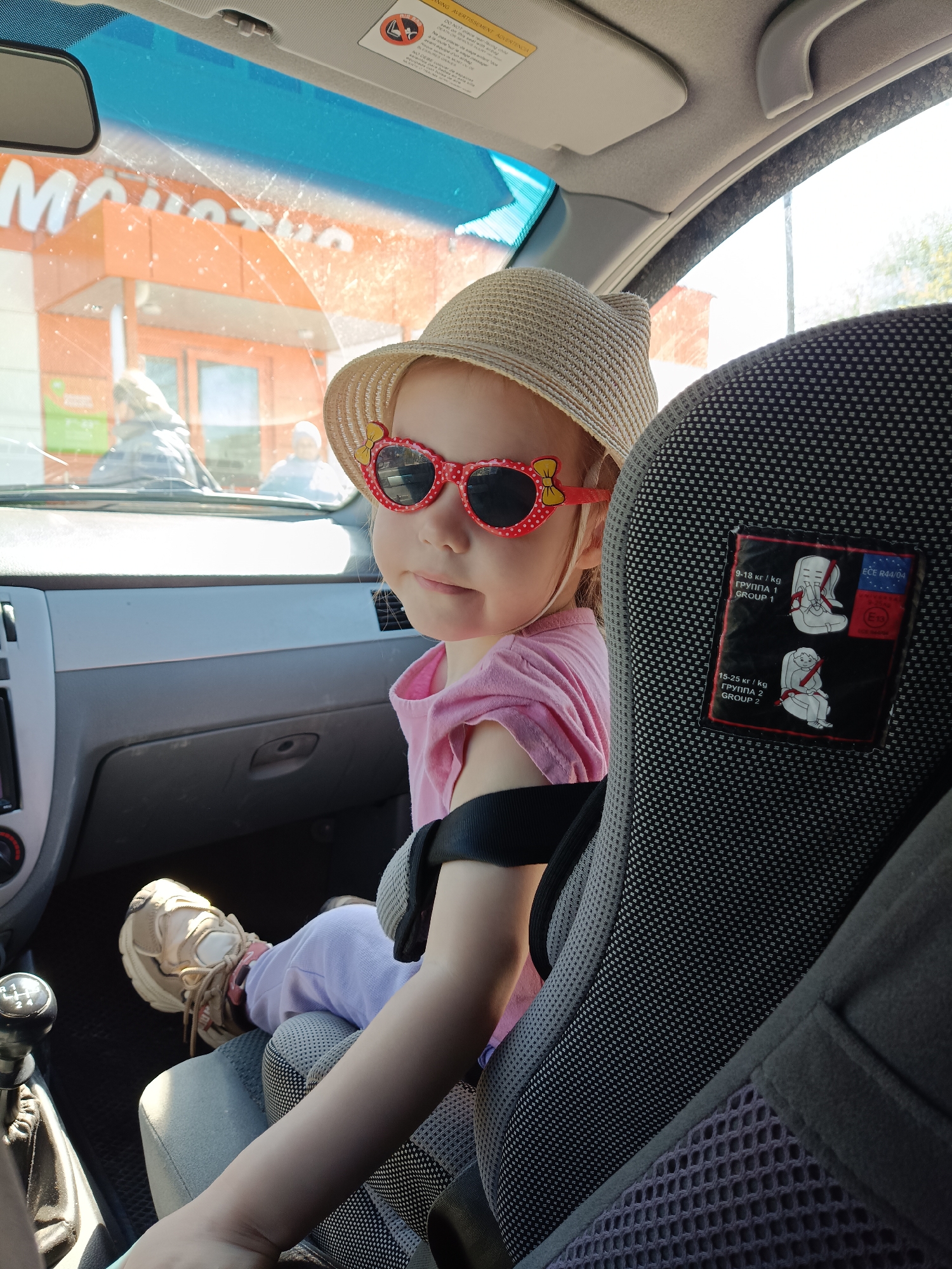 A young child is sitting in a car seat in the backseat of a car. The child is wearing a pink t-shirt, light purple pants, a beige straw hat, and sunglasses with red frames in the shape of hearts. The child is facing forward and looking to the left. The car seat is gray and black, with a label showing how to install the car seat for different weight classes. The child is wearing sneakers. In the background, a storefront with an orange facade and a sign displaying "MONETKA" in white letters can be seen. This suggests the location is near a Moneta store, likely in Ust-Kishert, Russia. The car is parked, and sunlight is coming in through the windshield. A gear shift is visible to the left of the child.