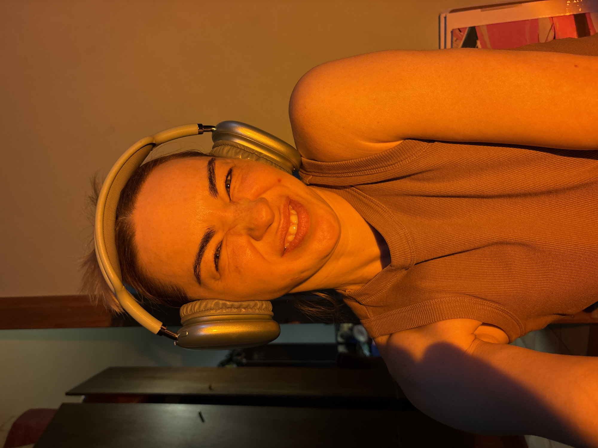 A woman with light skin and brown hair is wearing headphones and smiling widely. She is wearing a brown tank top. The lighting is warm, suggesting indoor lighting. The location is unknown, and there is no evidence the image was taken in Russia.