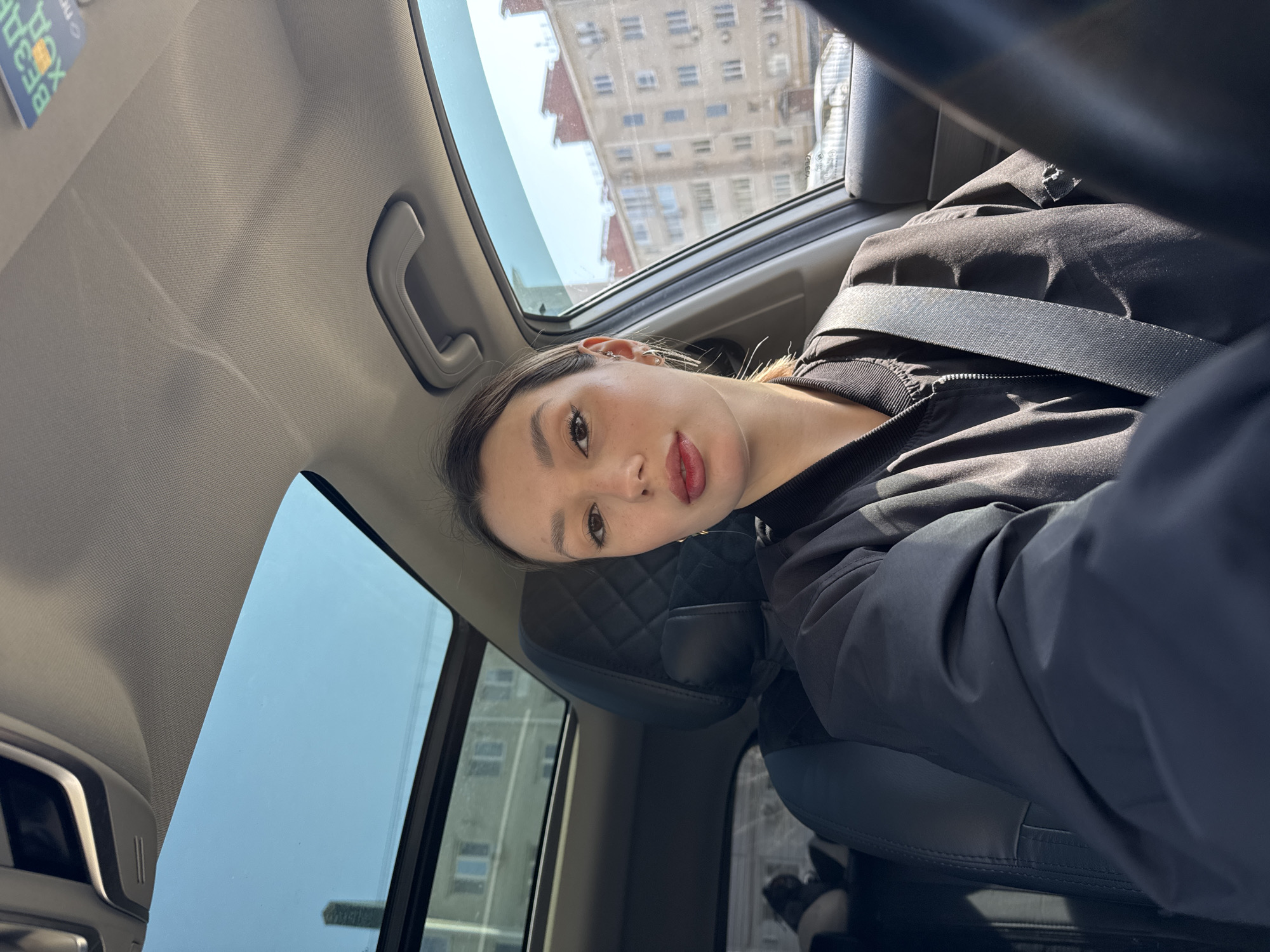 The image is a selfie of a young woman taken from a low angle inside a car. The woman, who appears to be of East Asian or mixed ethnicity, has dark hair, dark eyes, and is wearing red/pink lipstick. Her head is tilted as she looks directly into the camera. She is dressed in a black bomber-style jacket, with a car seatbelt fastened across her chest. Her ear is adorned with several small earrings and an ear cuff.
The car's interior features a light grey headliner with a grab handle visible on the right. The seats are dark, with the visible passenger seat headrest showing a quilted pattern. Outside the car windows, an urban scene unfolds with multistory residential buildings and a clear blue sky, indicating bright, sunny daytime weather. The sunlight creates distinct shadows within the car.
In the upper left corner of the car's headliner, an upside-down card or sticker is partially visible with text that includes "BESB3RA" and "XOP." The woman's expression is neutral, with a slight pout on her lips.