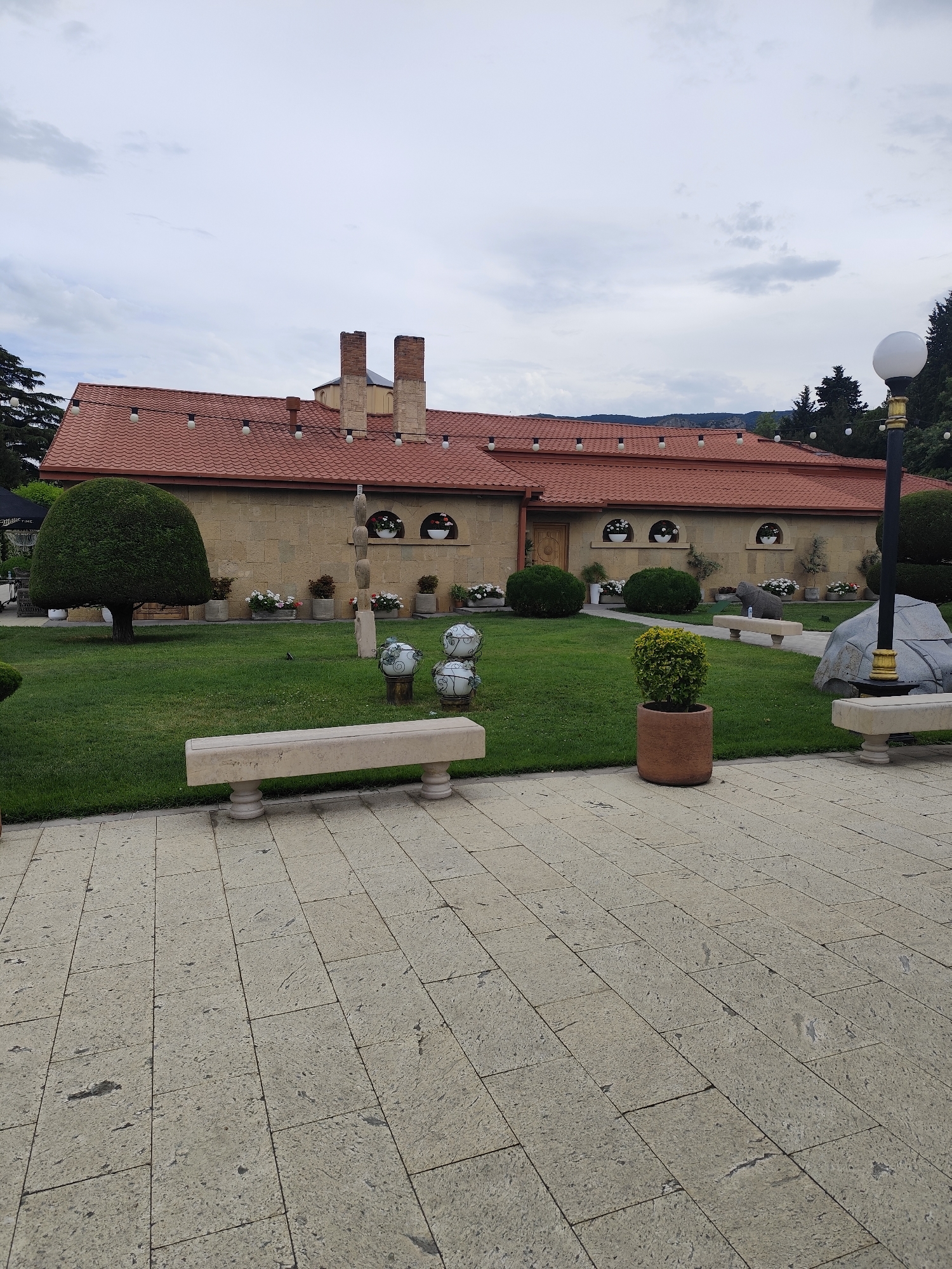 The image shows a courtyard in front of a building with a red tile roof. The building is made of light-colored stone and has two chimneys. In front of the building, there is a well-maintained lawn with several potted plants and decorative spherical sculptures on pedestals. A stone bench is placed on the lawn. The courtyard itself is paved with large, light-colored stone tiles. A lamppost stands to the right of the image. In the background, there are trees and hills, suggesting a semi-rural or park-like setting. The sky is overcast, indicating a cloudy day. There is no visible activity or people in the scene, giving it a peaceful and serene feel. The location is identified as Mtskheta, Georgia.