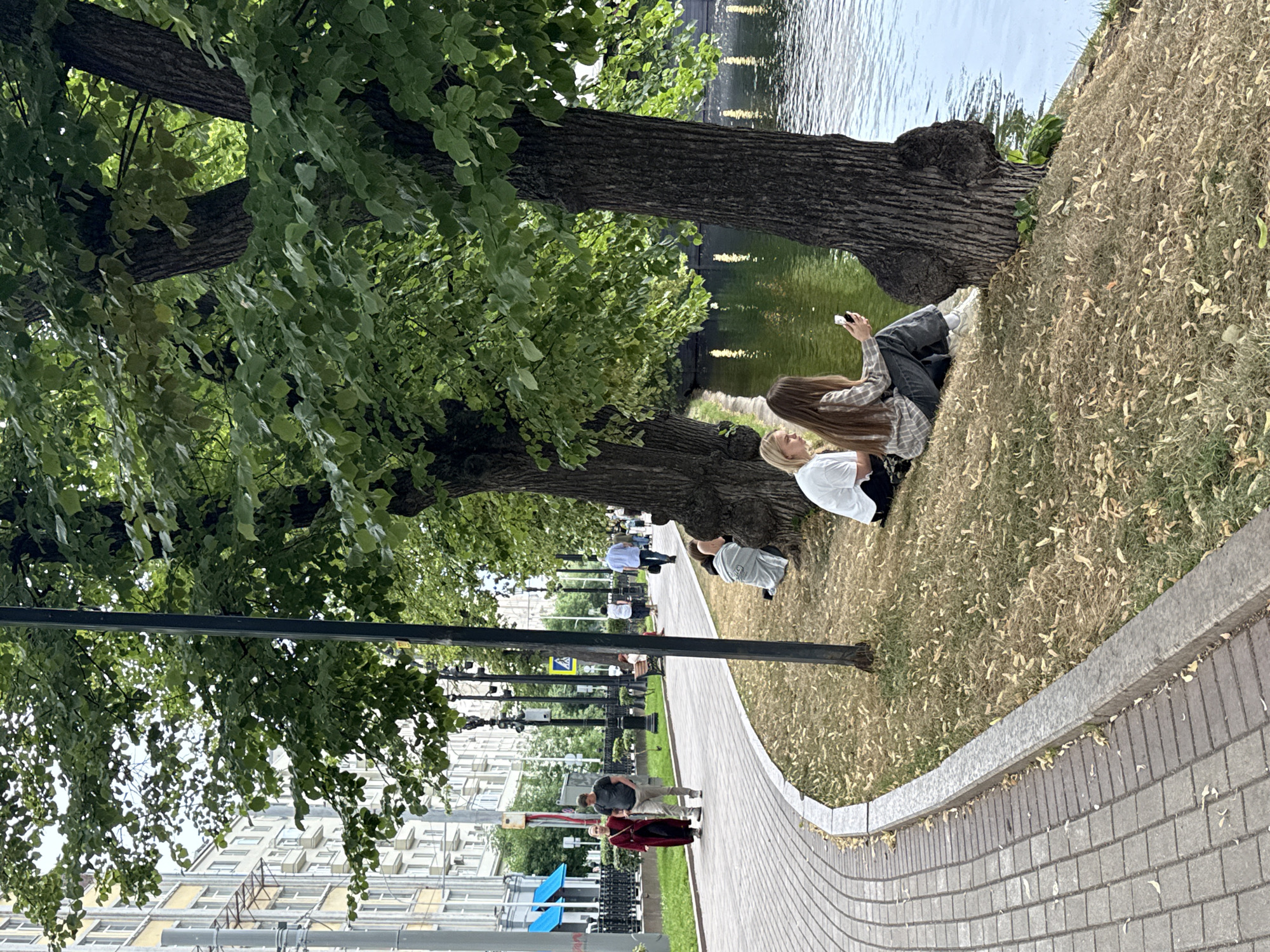 The image depicts a waterside promenade in Moscow, Russia, likely in the park area along the Moscow River or one of its canals. The scene is characterized by a paved walkway curving along the left side of the frame, bordered by a grey stone curb. Beyond the curb, a grassy area slopes down towards the water, covered with dry leaves. Several large trees with dense green foliage line the bank, providing shade.
In the foreground, two young women are seated on the grassy bank near the base of a large tree. One woman, with blonde hair, is wearing a white t-shirt and black bottoms. The other, with long brown hair, is dressed in a plaid shirt and dark jeans, holding a smartphone in her hand.
Further along the promenade, a couple is walking. The man is wearing a dark t-shirt and light-colored pants, while the woman is in a long, dark red dress. Other pedestrians are visible in the background, including individuals on benches and walking further down the path. Behind the promenade, a multi-story apartment building with yellow and white facades stands, indicating an urban setting. The water appears calm with reflections of light sources on its surface. A dark lamppost is positioned on the grassy area between the walkway and the trees.