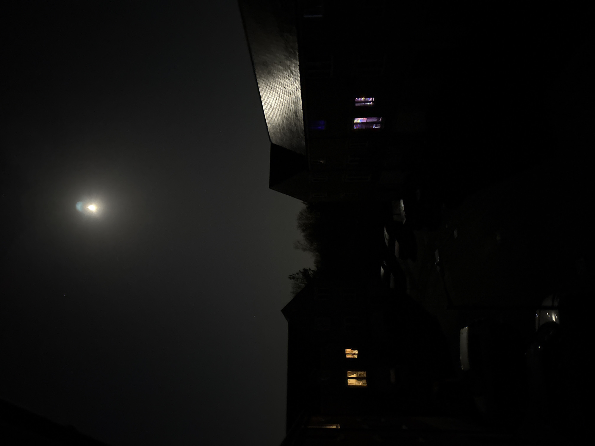 At night, a dimly lit urban scene in Great Yarmouth, United Kingdom. The sky shows a pale moon. A dark, rectangular building with a textured roofline extends from the upper left corner towards the center. To the right, a taller building with several illuminated windows with a variety of colors and patterns of light suggesting different uses. Below, a silhouette of a house with at least two windows showing warm, yellow light. The overall scene is very dark with the light sources providing the only visible detail.