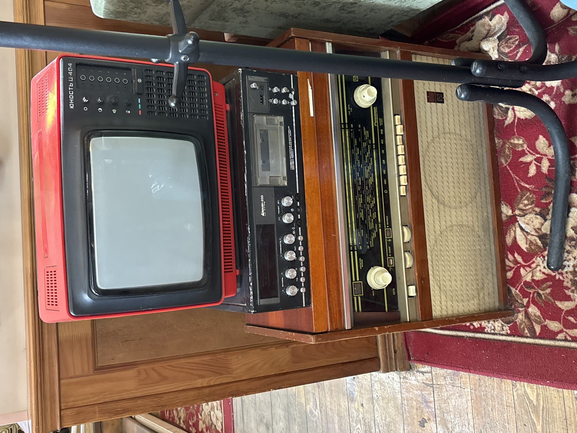 A cluttered indoor scene features a red Юность Ц-404 television, a black cassette player, and a "Рапсодия" radio, arranged on a wooden surface near a red and brown patterned rug. The scene appears to be located within a residential setting, with a glimpse of a door and a partially visible piece of furniture in the background. The precise location in Moscow, Russia, cannot be determined from the given information. The presence of a black metal structure over the items suggests that the arrangement might be for display or support purposes.
