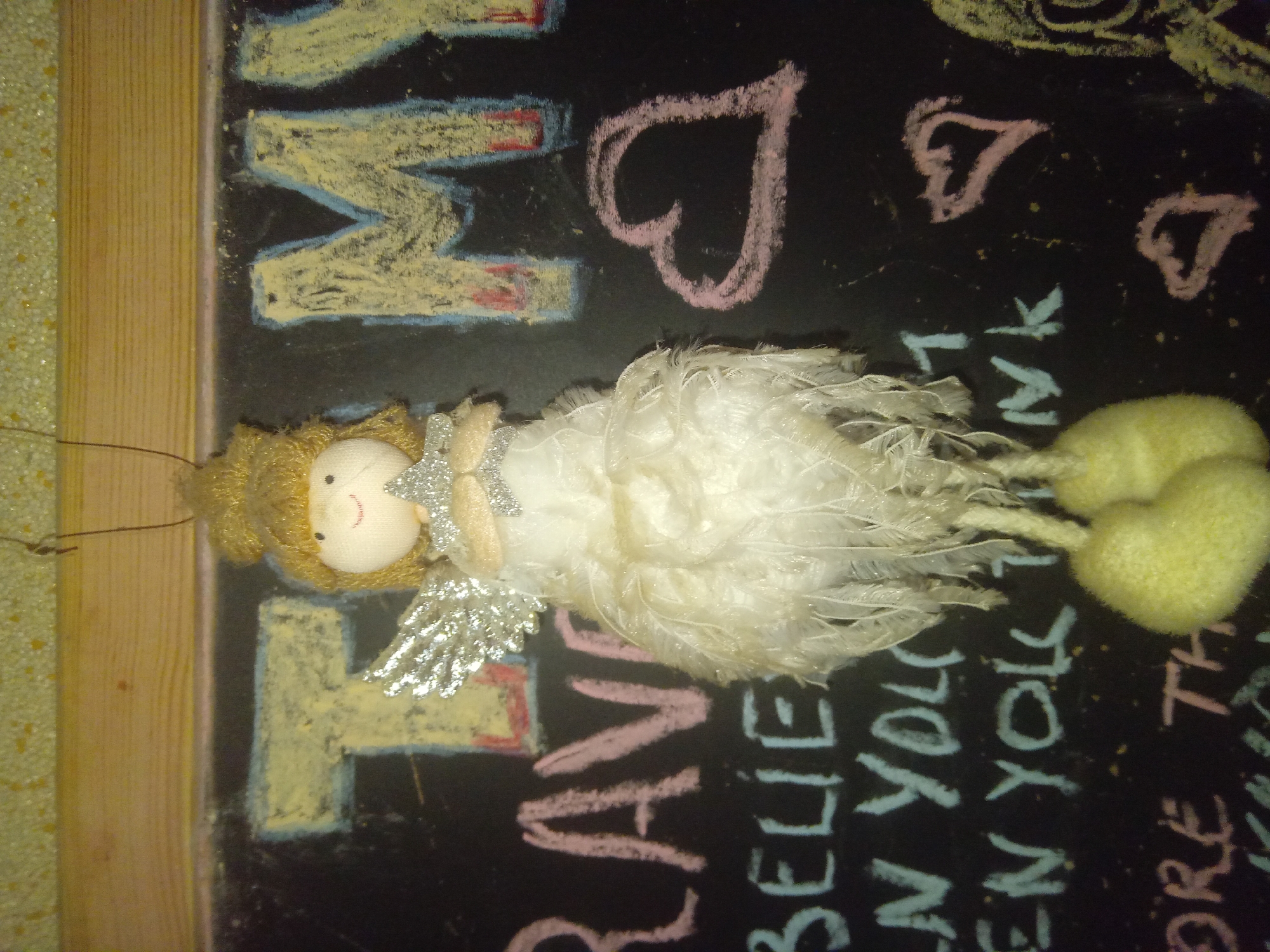 An overhead shot of a decorative angel ornament hanging in front of a chalkboard that has been drawn on with chalk. The angel is white with a yellow head and has a silver star in its hands. The chalkboard has the words "T" "MY", "RAVE", "BELIEVE", "IN YOU", "EN YOU", "nk", "ORE", "TH", and several heart drawings. The chalk writings appear to be motivational or inspirational quotes. The background of the image is out of focus but appears to be a wall with a yellow speckled texture. There is a wooden frame around the top and right sides of the chalkboard. The image was taken in Terebovlia, Ukraine.