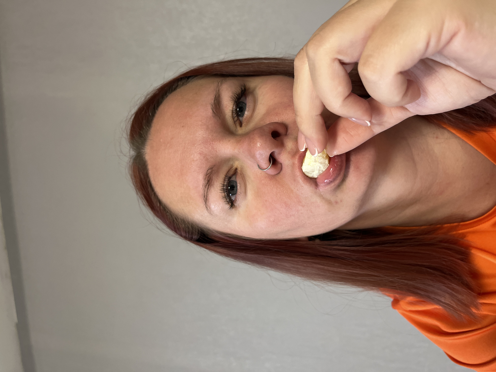 A woman with reddish-brown hair and blue eyes is eating. She is holding a piece of peeled fruit (possibly a tangerine section) between her fingers and placing it in her mouth. She has a silver nose ring in her left nostril. She is wearing an orange top. The background is a plain, light gray wall. The location is not discernible as Chaikovsky, Russia.