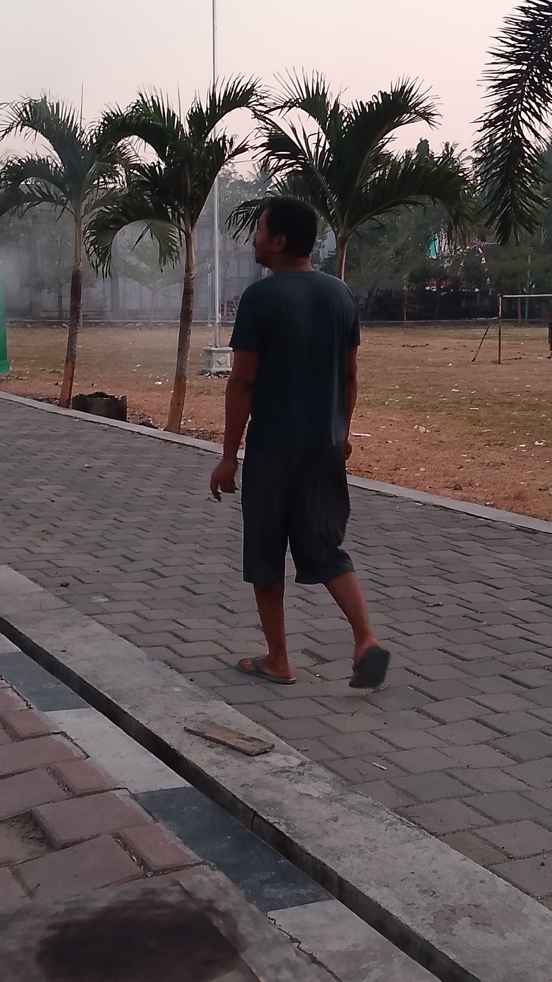 The image shows a full-body shot of a man walking away from the camera on a paved pathway. He is wearing a dark t-shirt and dark shorts, and flip-flops. The pathway is made of grey interlocking bricks. To the left of the pathway, there is a concrete curb and then a patch of grass. Beyond the grass, there are several palm trees and a sandy area, possibly a sports field. In the background, there are more trees and a hazy sky. The overall scene appears to be an outdoor public space, likely a park or recreational area, in Karanglangu, Indonesia.