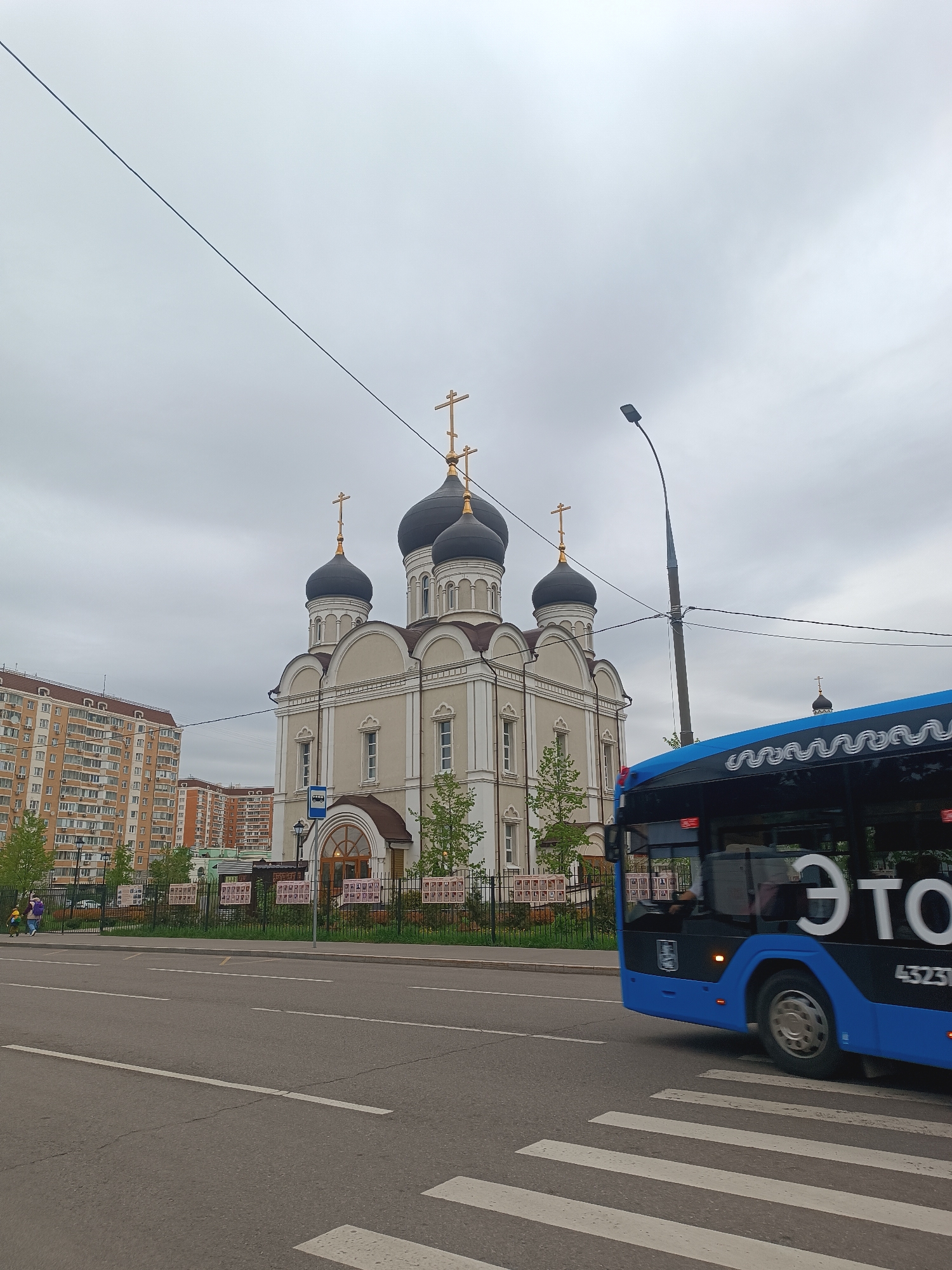 The image shows a street scene in Moscow, Russia, with a Russian Orthodox church in the background. The church has multiple black domes topped with golden crosses. To the left of the church are apartment buildings. A blue bus is passing in the foreground, partially obscuring the view. The sky is overcast and the road is marked with a zebra crossing. A few people can be seen walking on the sidewalk near the church. There are also signs along the fence surrounding the church.