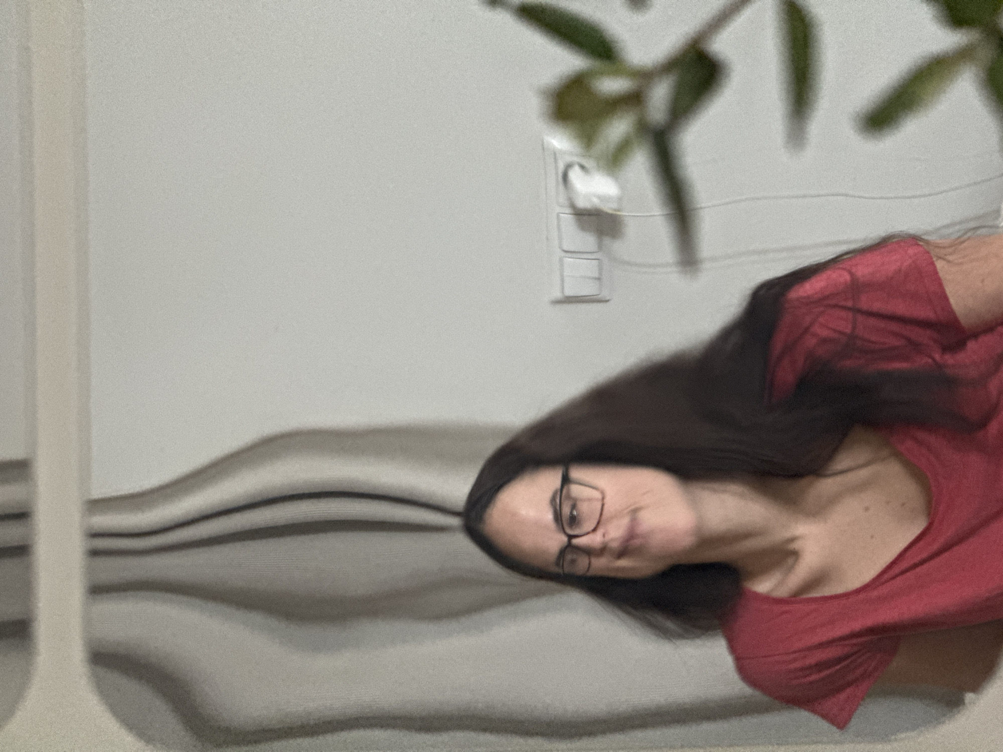 A woman with dark hair and glasses, wearing a red t-shirt, is shown in the image. Behind her is a white wall with a light switch and a plant. There's also a piece of furniture with a gray, wave-like pattern. There is no definitive information indicating the specific location in Krasnoyarsk, Russia.