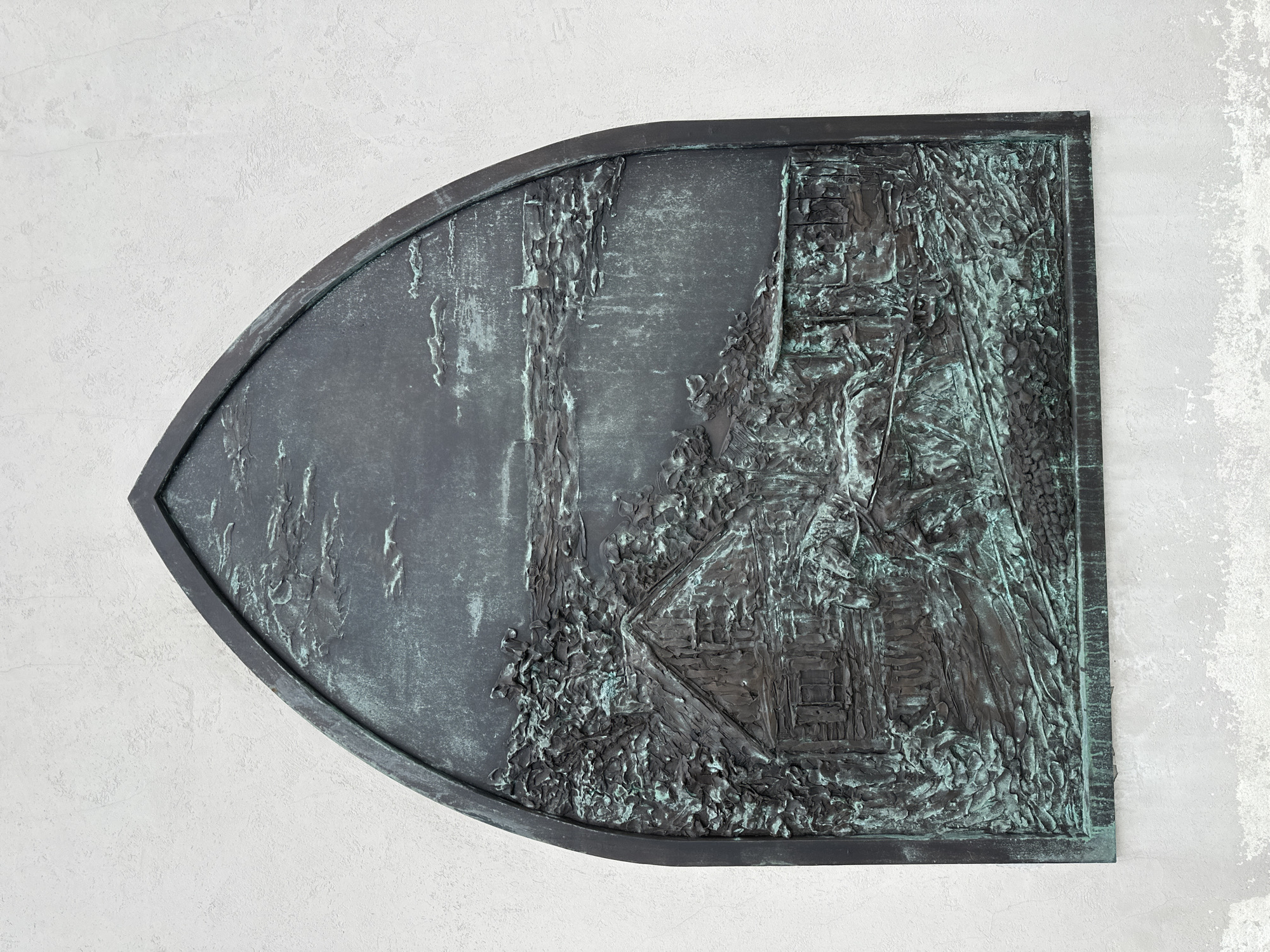 A bas-relief sculpture, framed by a pointed arch, is mounted on a white, textured wall. The sculpture depicts a rural scene with a wooden house in the left foreground, surrounded by dense foliage. To the right of the house, a horse-drawn cart is visible. In the background, a body of water stretches towards a distant, indistinct shoreline with structures. The sculpture's surface exhibits significant verdigris patina, indicating age and exposure. The overall impression is one of a historical or commemorative piece, likely depicting a specific place or event. This bas-relief is located on the facade of the National Library of Belarus in Minsk, Belarus.