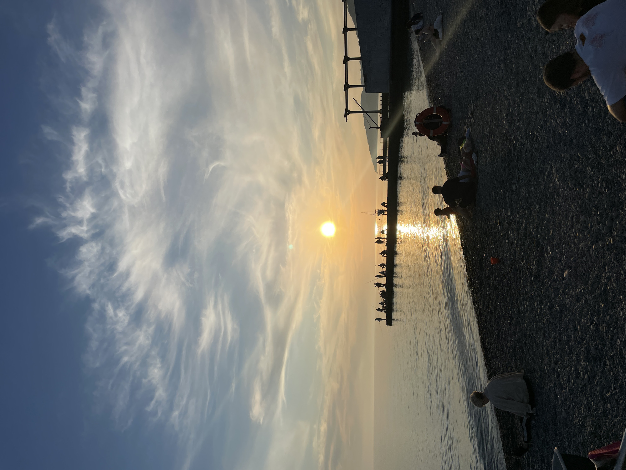 This image depicts a sunset scene on a pebble beach in Sochi, Russia. The sun, a bright golden orb, is low on the horizon, casting a strong reflection across the calm sea.
In the midground, a dark concrete pier extends into the water. Approximately 15 to 20 individuals are silhouetted on the pier, many appearing to be fishing with rods extending over the water. The sun's reflection illuminates the water directly in front of the pier.
The sky exhibits a clear blue gradient transitioning to lighter hues near the horizon, with distinct white cirrus and cirrocumulus clouds scattered throughout.
The foreground is dominated by a dark grey pebble beach. Several groups and individuals are present. On the far left, a person with light hair and a light-colored top sits facing the water. In the mid-right, a red lifebuoy is mounted on a stand. Near it, an adult male silhouette sits with a small child close to the water's edge. Further back on the beach, other figures are seated, some on blankets. In the bottom right corner, two individuals, an adult male in a white shirt and a female, are seated with their backs to the viewer. An orange bucket is visible on the pebbles to the left of these groups.
On the right side of the image, behind the pier, a low, light-colored concrete structure with a dark open-frame pergola is visible. Distant hills or mountains are silhouetted against the horizon in the far right background.