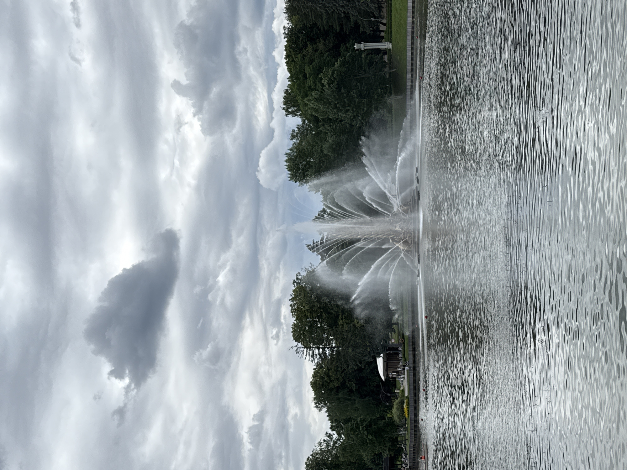 This is a photograph of a large, ornate fountain in operation on a lake, likely in a park setting. The fountain is centrally located, with water spraying high into the air in a fan-like pattern. The lake's surface is rippled, reflecting the overcast sky and surrounding trees. In the background, a line of lush green trees forms a dense treeline. On the shore, there are some park structures, including a white pavilion-like building and possibly some benches or seating areas where a few people are visible. A white columned structure is also visible on the right side of the image. The sky is filled with dramatic, dark grey clouds.
This scene depicts the Samson Fountain in Moscow's VDNKh (Exhibition of Achievements of National Economy) park. The fountain is a prominent landmark in the park and a popular tourist attraction. The VDNKh is a large exhibition center and park located in the northeast of Moscow, Russia.