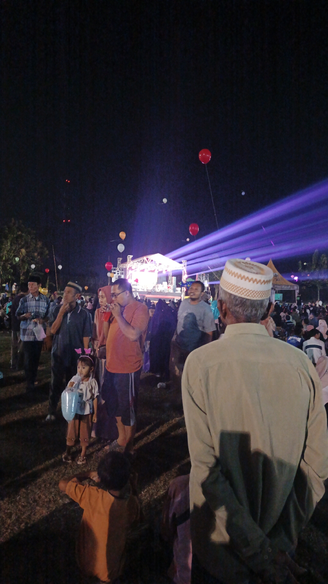 In Ketemasdungus, Indonesia, an outdoor evening event is taking place. A crowd of people, many of whom are wearing traditional Islamic attire like peci caps and hijabs, are gathered on a grassy field. A stage with bright lights and what appears to be a performance or presentation is visible in the background. Above the crowd, several red, white, and orange balloons are floating in the dark sky, some illuminated by the stage lights. The overall atmosphere suggests a festive or celebratory occasion, possibly a religious or cultural gathering.