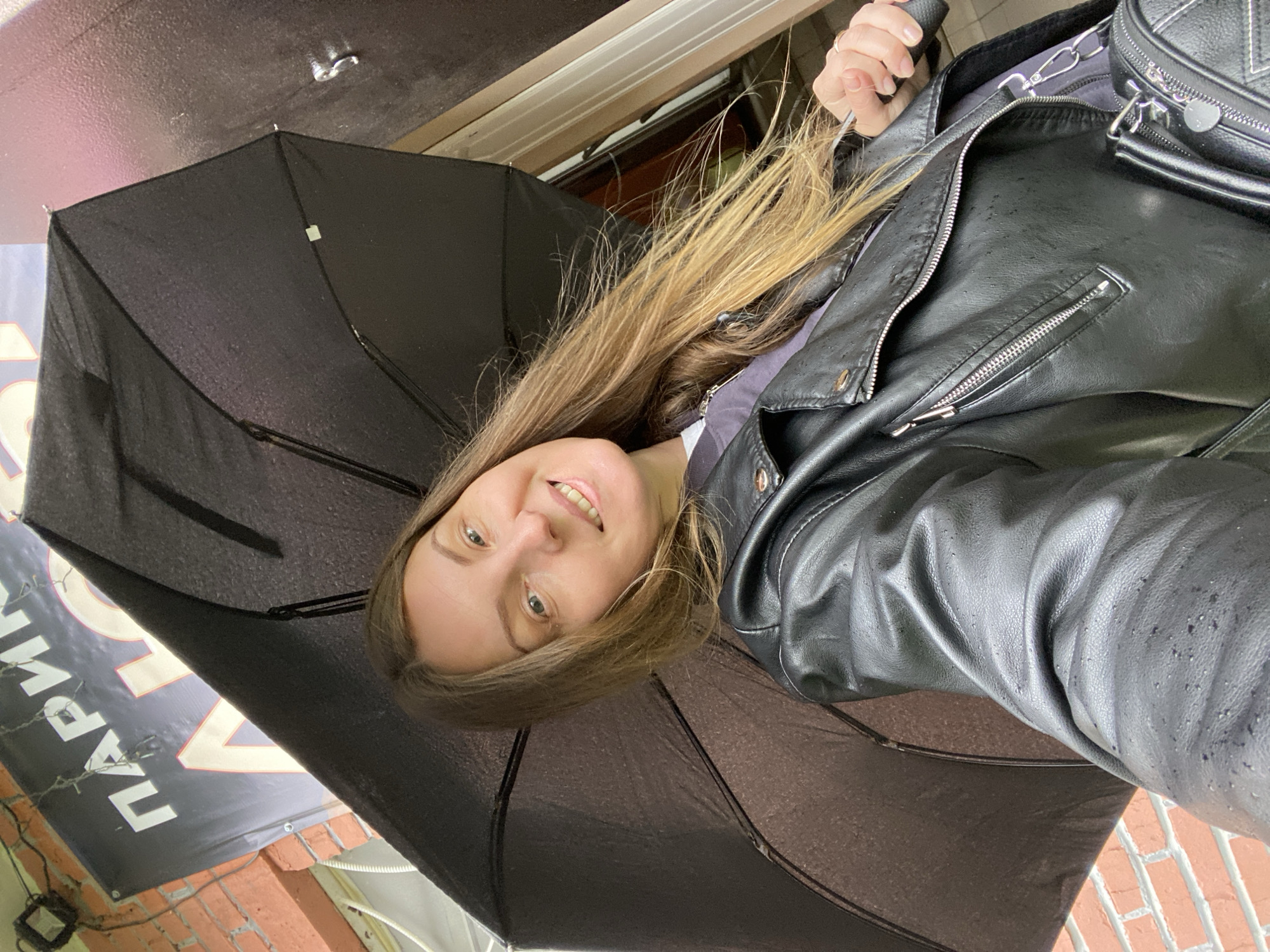A woman stands under a black umbrella in Noviy, Russia. She is wearing a black leather jacket. The scene is outdoors and appears to be overcast or raining. A sign with Cyrillic script and red brickwork are visible in the background.