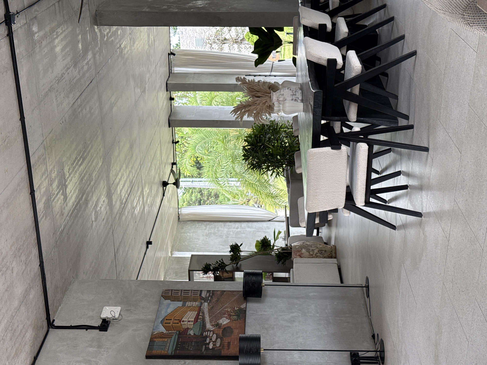 The image depicts an indoor setting in Ubud District, Indonesia. The interior is characterized by a concrete structure with large windows overlooking lush greenery. A long table is present with a row of chairs on one side, facing the windows. The chairs have a black frame and a light-colored fabric seat and back. The table is decorated with potted plants and vases. A painting is visible on the wall opposite the windows, illuminated by a black track lighting system. The flooring appears to be tiled. There are no people present in the scene. The environment is bright, with natural light streaming through the windows, highlighting the vegetation outside.