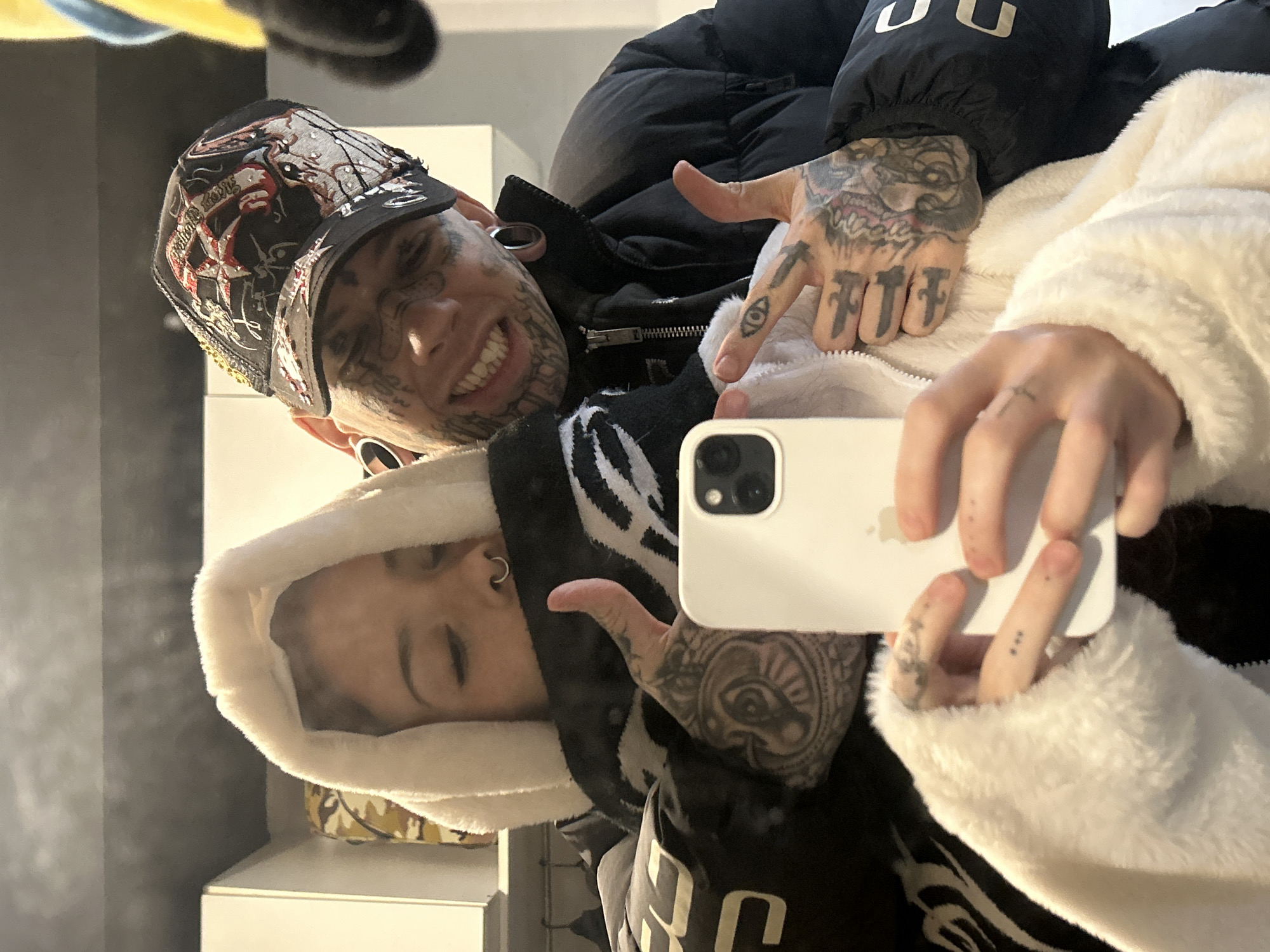 A selfie shows two individuals. One individual, positioned to the left and partially in the foreground, displays extensive facial and hand tattoos and is wearing a hat. The other individual, slightly behind and to the right, wears a hooded garment. Both individuals are holding a white smartphone, presumably taking the selfie. The background is out of focus, but appears to include a wall, shelves, and other objects, suggesting an interior space. The location in the city of Buenos Aires, Argentina is unknown.