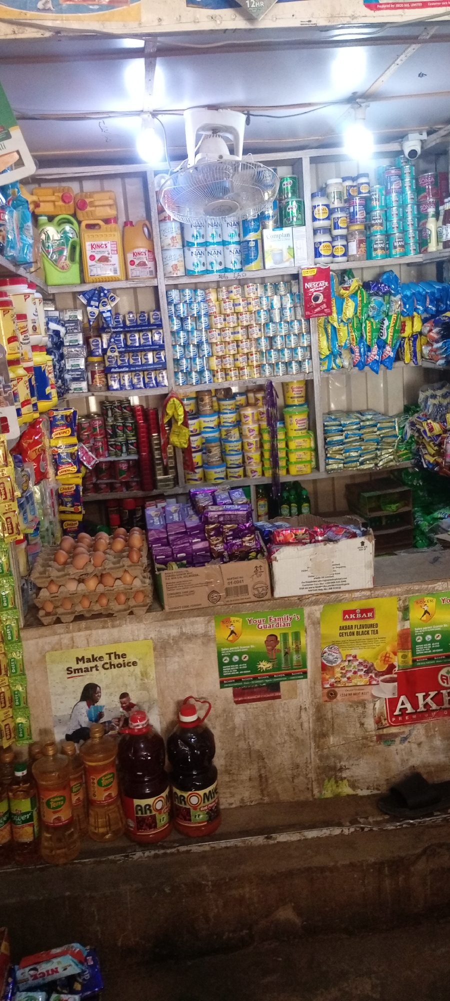 The image captures the interior of a small retail shop in Aviation Quarters, Nigeria. Shelves are densely stocked with a variety of goods, including: eggs, oil bottles, bottled juice, baby formula (NAN, Complan), canned food, milk products (Hollandia), cocoa beverage (Nestle), sweets, detergent, and tea (AKBAR). The lighting is provided by a combination of fluorescent tubes and a ceiling fan with integrated light. Advertisements and posters are visible on the walls, including brands such as AKBAR tea, Boxer insecticide and a campaign promoting healthy food choices. A security camera is mounted near the top shelf. A pair of sandals is placed on the right edge of the image.
