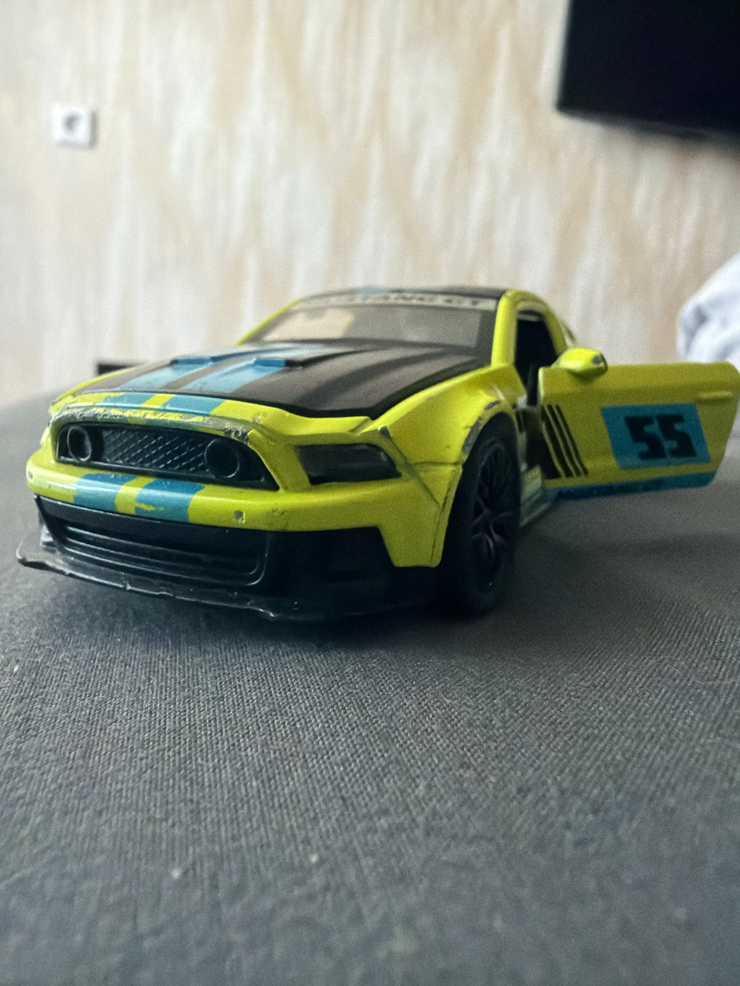 A bright yellow toy car with blue and black racing stripes is positioned on a textured gray surface, possibly a couch or fabric-covered table. The car is a replica of a Ford Mustang, evident from its distinctive grille and body shape. The driver's side door is open, revealing the interior. A blue rectangular decal with the number "55" in black is visible on the door. The car has black wheels and a black hood with blue racing stripes. In the background, a textured beige wall with a small square electrical outlet is visible. The lighting suggests an indoor setting. The image lacks any human presence and there are no discernible indicators of its specific location within the city of Hrodna, Belarus.