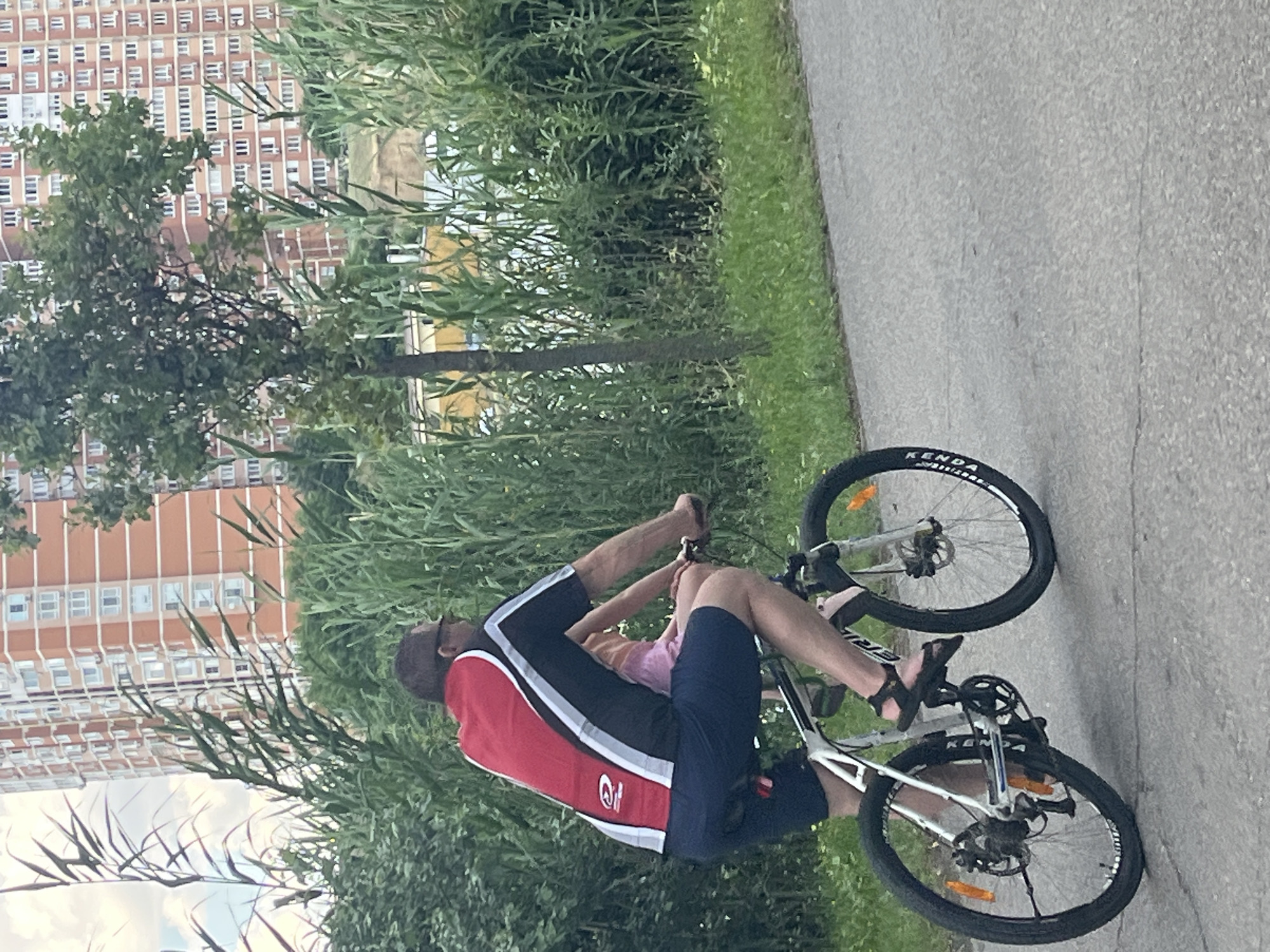 A man is riding a bicycle with a young child sitting on his lap. The man is wearing a red, black, and white cycling jersey and dark blue shorts. He has on dark sandals. The child is wearing a light pink top and light-colored shorts. The bicycle is a mountain bike with black tires. The scene appears to be a park or recreational area, with tall green grass and trees on the left side of the frame. In the background, there is a large, multi-story apartment building with many windows. The asphalt path is on the right side of the image. The lighting suggests it is daytime, possibly afternoon, with overcast skies. The image has a casual, everyday feel. There is visible text on the bicycle tire that reads "KENDA". The location context is Krasnodar, Russia.