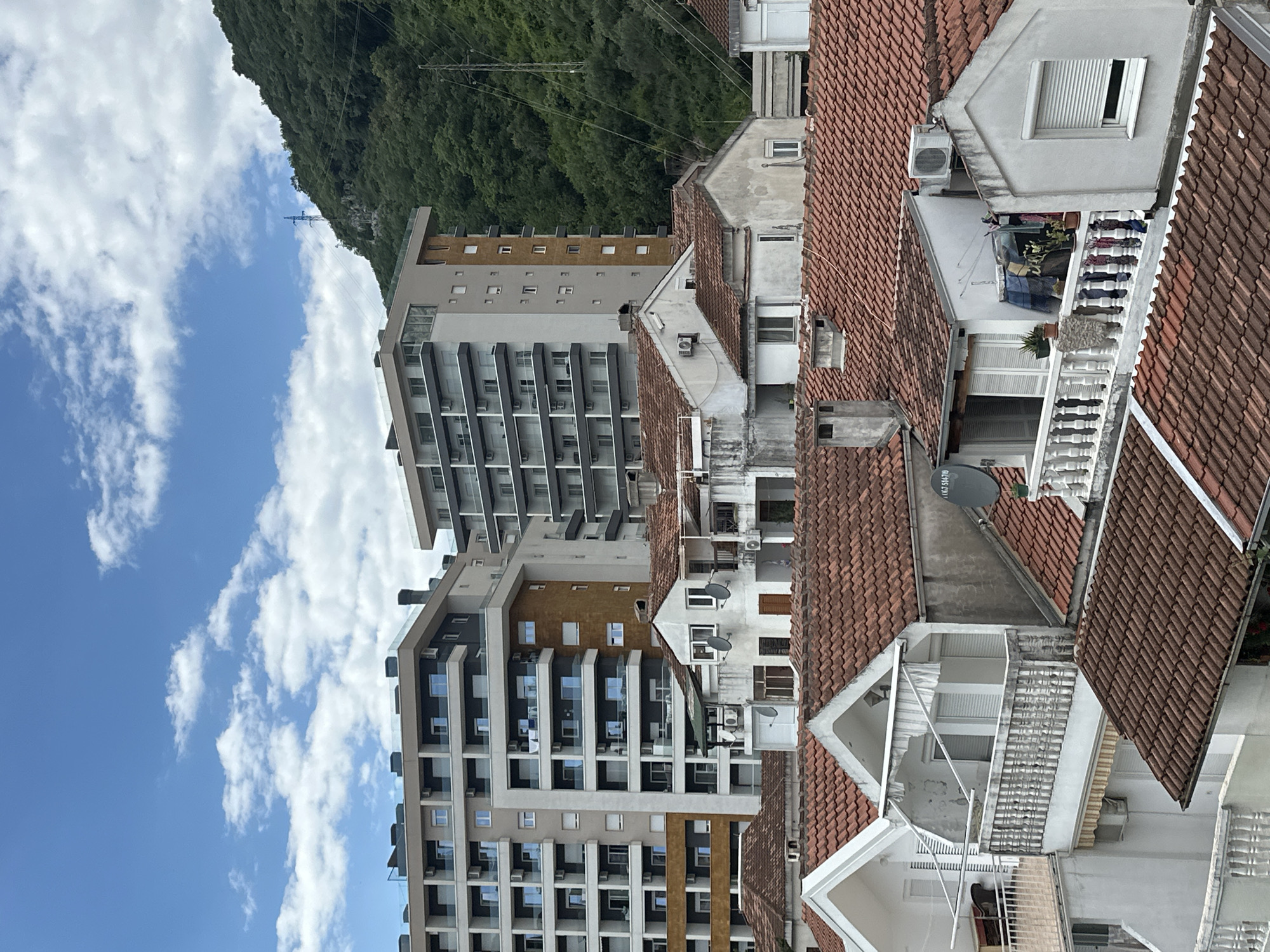 The image shows a view of buildings in Budva, Montenegro. A modern high-rise apartment building, with multiple balconies and windows, stands next to older buildings with terracotta tiled roofs. Some of these older buildings feature white walls, balconies with ornate railings, and satellite dishes. In the background, a tree-covered hill rises toward the sky. The sky itself is partly cloudy with blue patches. Power lines are visible against the backdrop of the hill.