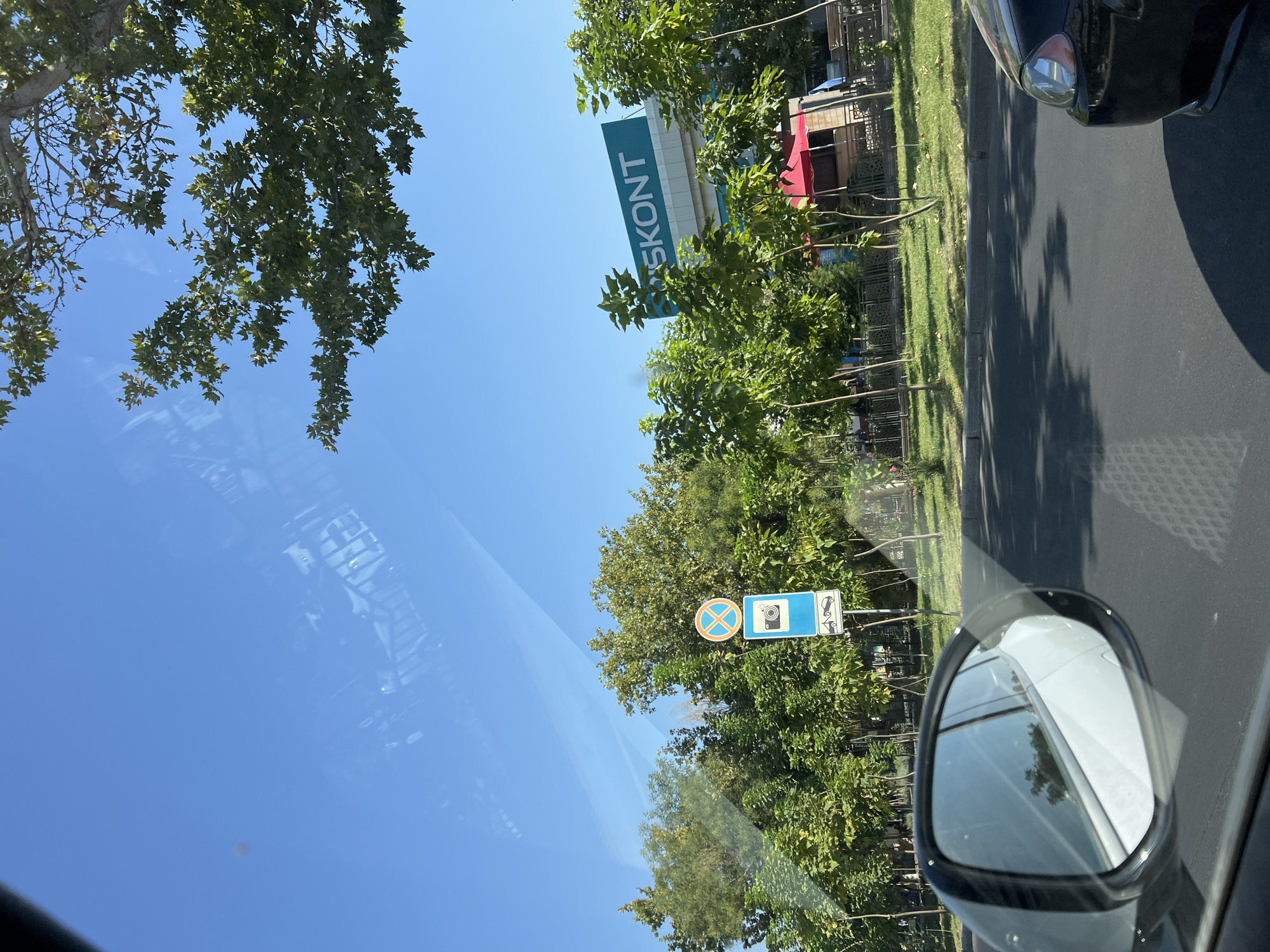 The image depicts a street scene viewed from a vehicle. In the foreground, the side mirror of a white car is visible on the left, reflecting trees and sky. The road is paved, with a grey curb separating it from a grassy area with young trees. On the right side of the image, the front headlight and fender of a black car are visible.
In the background, a building with a large teal sign that reads "SKONT" is prominent. Several trees with green foliage line the street, and a red awning can be seen over a structure in front of the building. There are also traffic signs visible; one sign shows a camera icon, indicating a speed or traffic camera, and another sign above it has a red circle with a diagonal line through it, suggesting a prohibition. The sky is clear and blue. There are no visible people in the image.