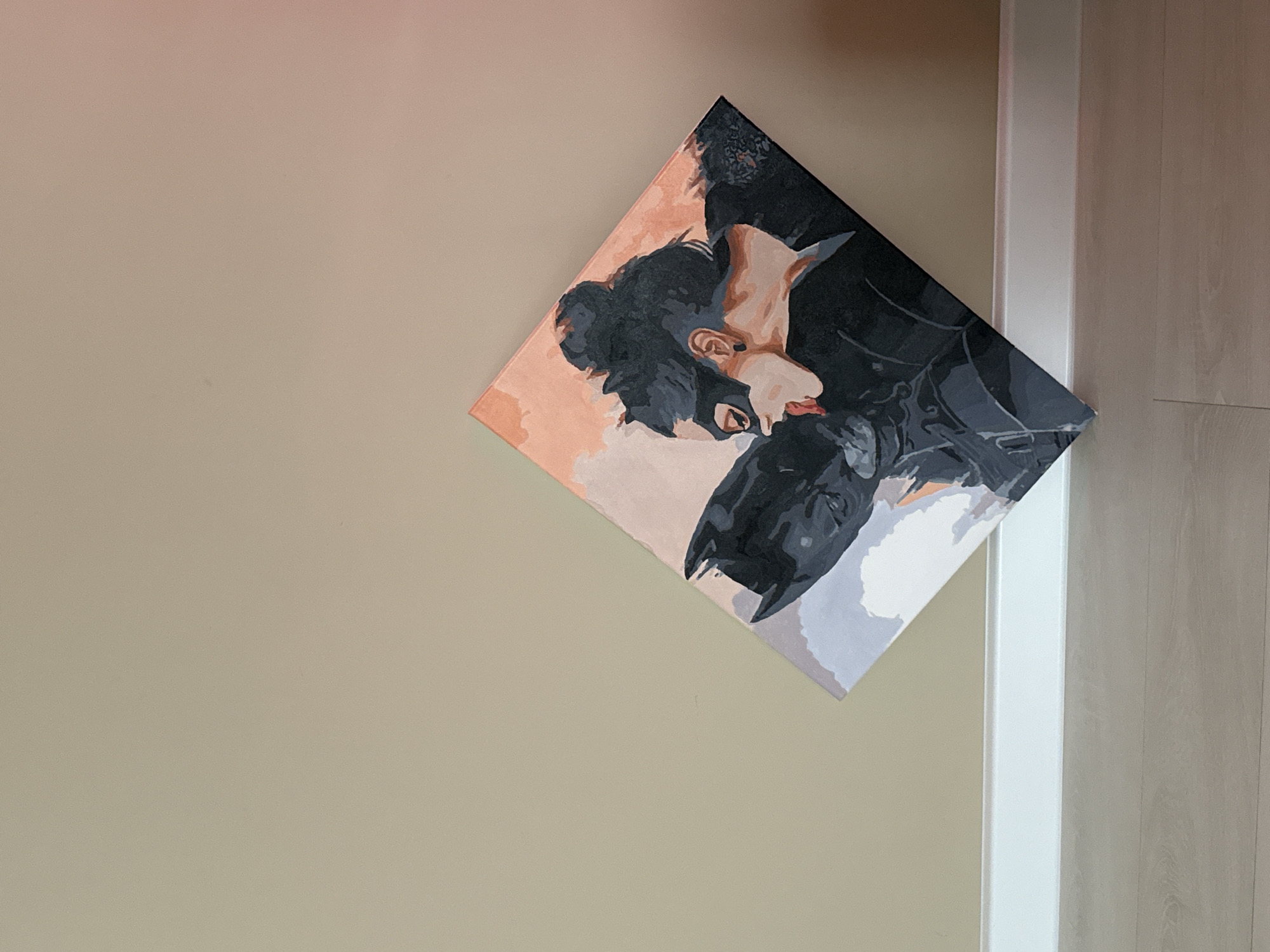 A square painting, oriented diagonally like a diamond, is mounted on a light beige wall. The artwork depicts two stylized figures in an intimate pose, rendered in a blocky, possibly paint-by-numbers, art style using shades of black, grey, peach, and light brown.
The figure on the left, likely Catwoman, has dark hair pulled into a bun and wears a black, cat-eared mask. Their face, rendered in warm skin tones, leans towards the second figure, with red lips slightly parted. The figure on the right is identifiable as Batman, wearing his classic black, pointy-eared cowl. Their faces are very close, suggesting a tender kiss or embrace. The background of the painting features abstract color blocks.
To the right of the painting, a white vertical door frame or trim is visible, leading to a section of light-colored wooden floor or wall paneling. The room appears to be indoors and well-lit. There is no visible text in the image.