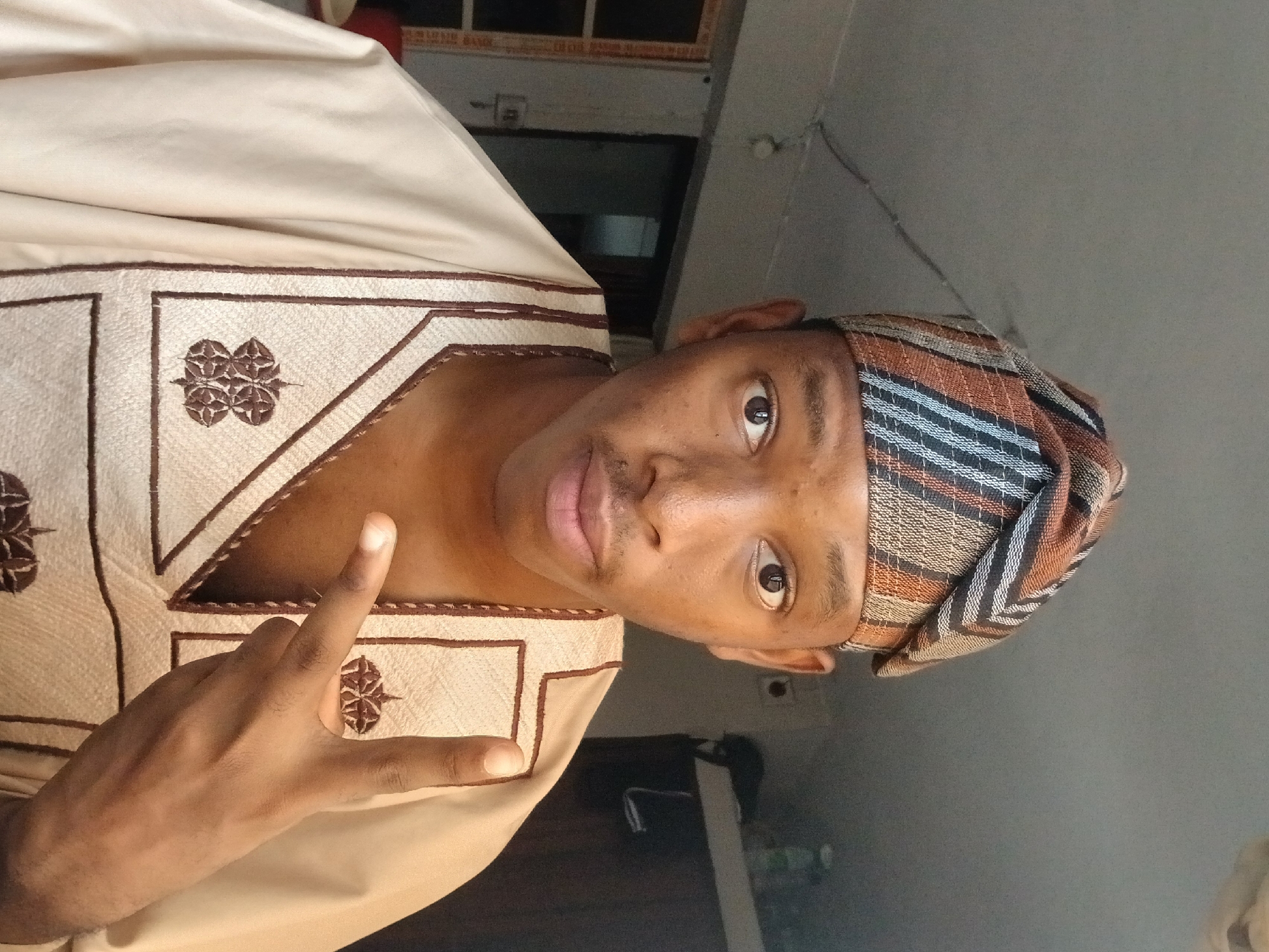 The image shows a young Black male wearing a traditional Nigerian outfit consisting of a light-colored tunic and a patterned headwrap. He is indoors, with a neutral-colored wall and ceiling visible. In the background, a doorway and a shelf with some items are discernible. The man is looking directly at the camera and making a hand gesture with two fingers extended. There is no discernible indication of the specific location within Lagos, Nigeria.