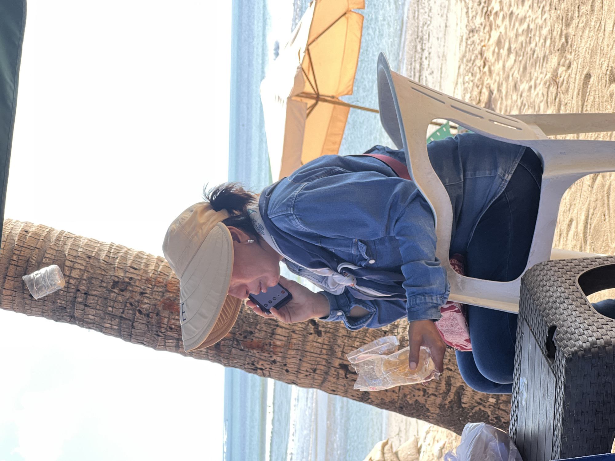 A woman sits on a white plastic chair on a sandy beach in Kuta, Indonesia, talking on her cell phone and holding a plastic bag with food. She is wearing a denim jacket, dark pants, a wide-brimmed hat, and a scarf. Behind her, a palm tree trunk is visible, with a plastic bottle attached to it. The ocean, with waves and a distant umbrella, is in the background. A woven plastic table is in the foreground.