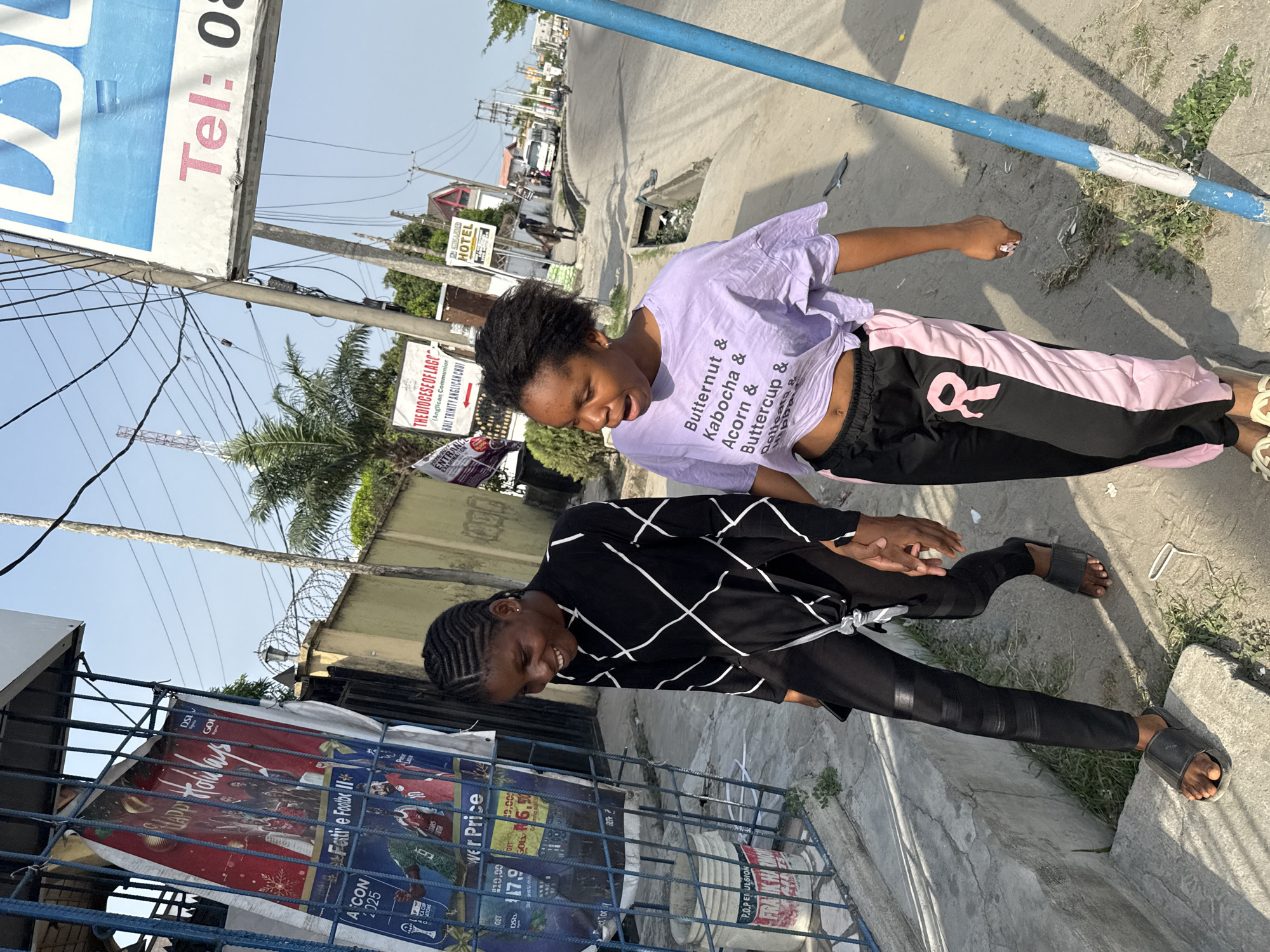 The image depicts two women standing close to each other outdoors. The woman on the right wears a light purple shirt with the words "Butternut & Kabocha & Acorn & Buttercup & Delicata & Hipba." She has black and pink sweatpants with a ribbon design and wears sandals. The woman on the left wears a black shirt with white stripes and black pants, also wearing sandals.
In the background, there is street signage including "Tel: 08", "THE DIOCESE OF LAGOS (Anglican Communion HOLY TRINITY ANGLICAN CHUF," and "HOTEL." A promotional banner displays "DS" and "GOI," along with "Festive Footbe II" and "ASCON 2025." The surrounding area suggests an urban setting with trees, buildings, and a visible road. The street is likely located in Ado, Nigeria.