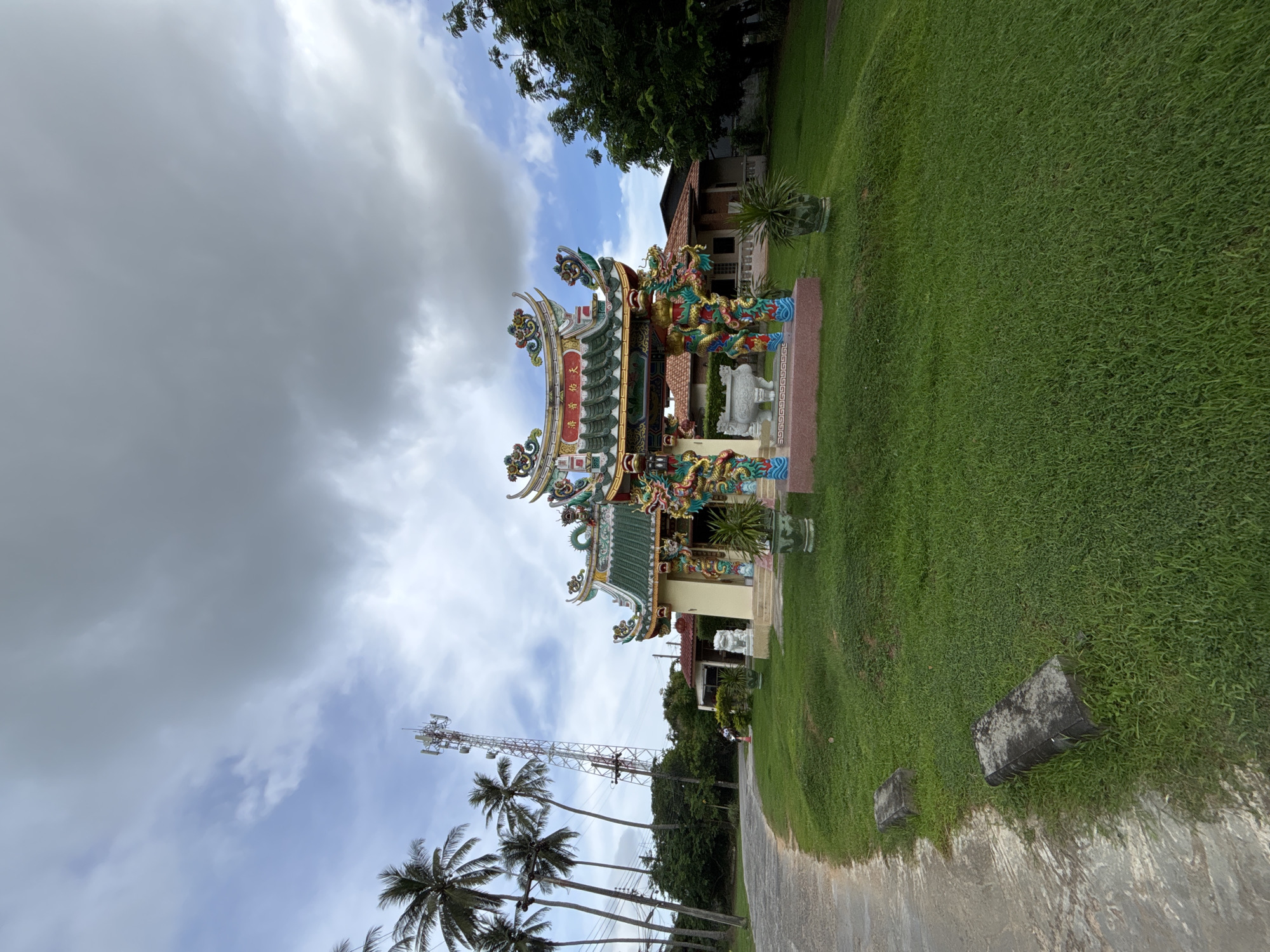 In Chalong, Thailand, a vibrant, ornate Chinese temple gate stands prominently on a well-manicured grassy knoll. The gate features intricate detailing, including colorful dragon sculptures and ornamental roof crests, with traditional Chinese characters adorning the upper facade. To the left of the temple, a paved pathway curves away, bordered by lush green grass and several large, weathered stone slabs. In the background, a tall telecommunications tower pierces the cloudy sky, and a row of tall palm trees lines the left side of the scene. Behind the temple, a single-story building with a terracotta roof is visible, partially obscured by foliage. The overall environment suggests a humid, tropical climate, typical of southern Thailand, under a sky that is partly overcast with dramatic grey clouds. There are no discernible people actively engaged in the scene.