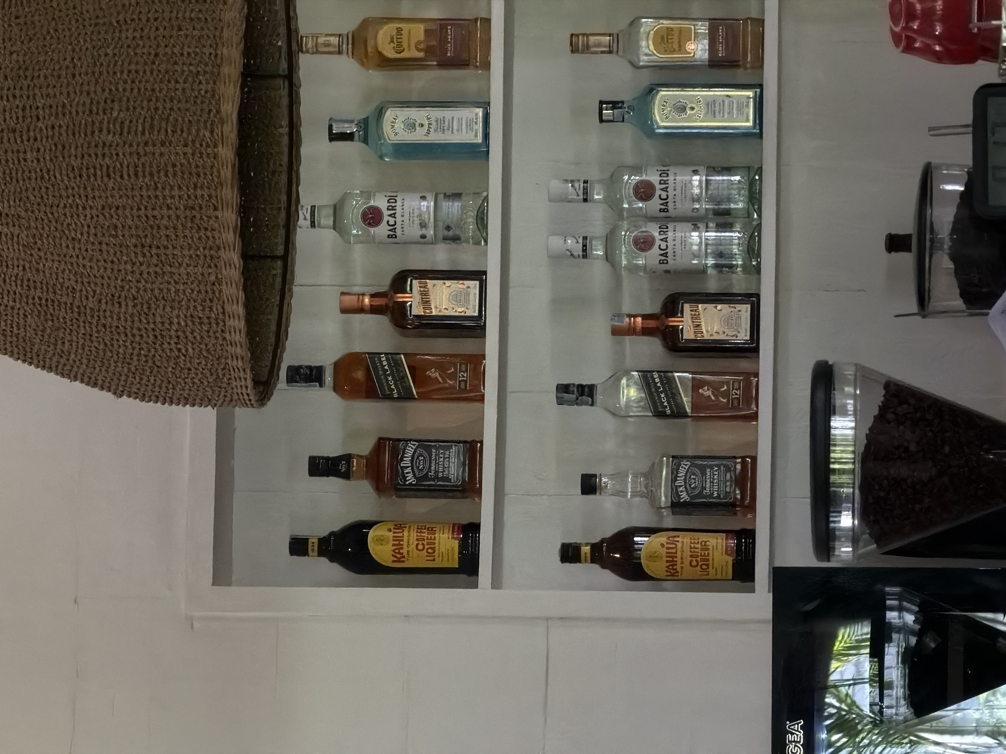 The image shows a shelf stocked with liquor bottles. Brands visible include Cuervo, Bombay Sapphire, Bacardi, Cointreau, Johnnie Walker Black Label, Jack Daniel's, and Kahlua. A container of coffee beans is also on the shelf. The image was taken in North Kuta, Indonesia.