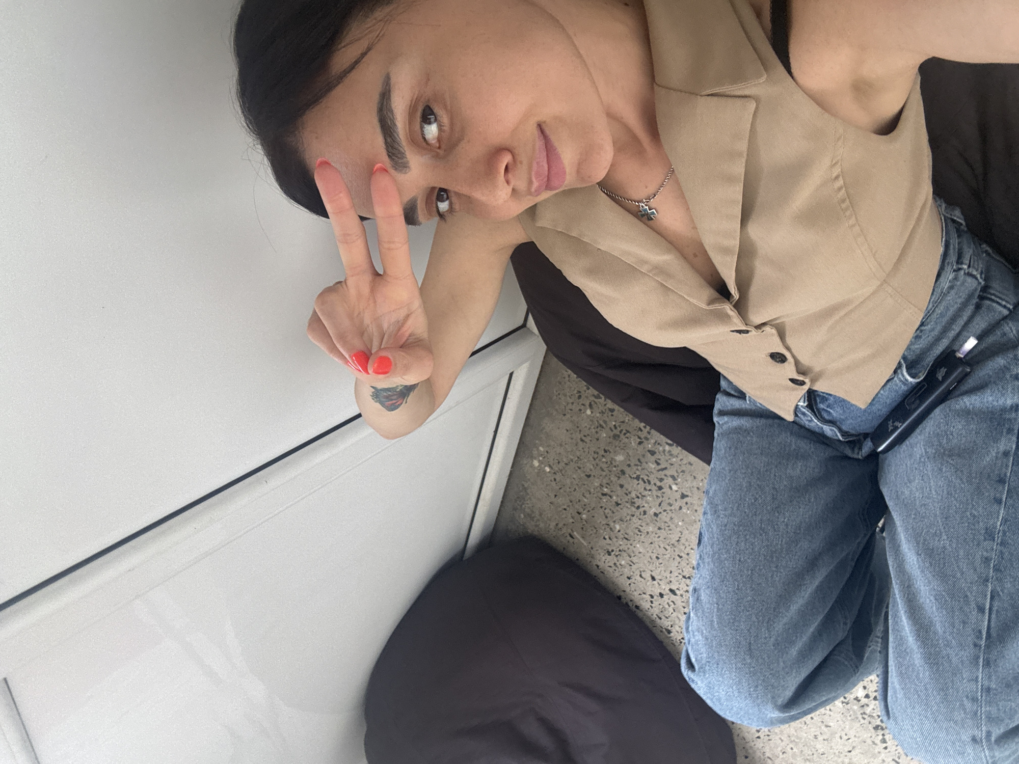 The image shows a woman with dark hair and fair skin, wearing blue jeans and a tan vest. She is sitting down, possibly on a beanbag chair, and has her right hand raised to her forehead with two fingers extended, forming a peace sign. Her nails are painted orange. She is wearing a silver necklace with a cross pendant. A black object, likely a phone, is visible in her lap. The background consists of a white wall or door and a speckled gray floor. The scene appears to be indoors, with no specific identifying markers of Tbilisi, Georgia.