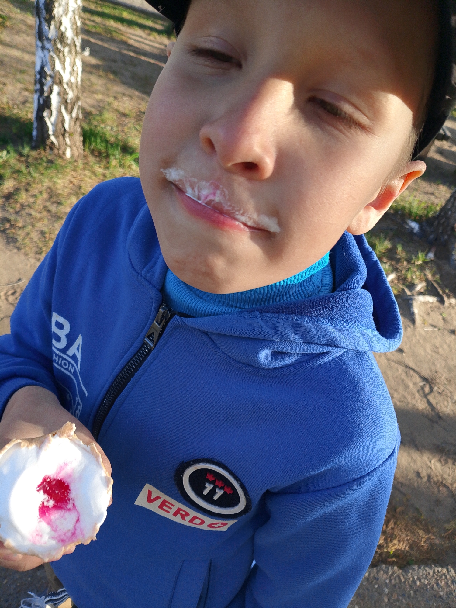 A young boy with fair skin and a milk mustache is holding a nearly-empty ice cream cone. He wears a blue hoodie with a logo patch that reads "VERD" and another patch with the letters "BA" on the sleeve. He has a blue turtleneck underneath the hoodie. The location is outdoors, likely in the city of Noviy, Russia, given the surrounding context. A tree with white bark is visible in the background, along with some ground and a paved area. The boy's eyes are mostly closed, and he appears to be enjoying his ice cream.
