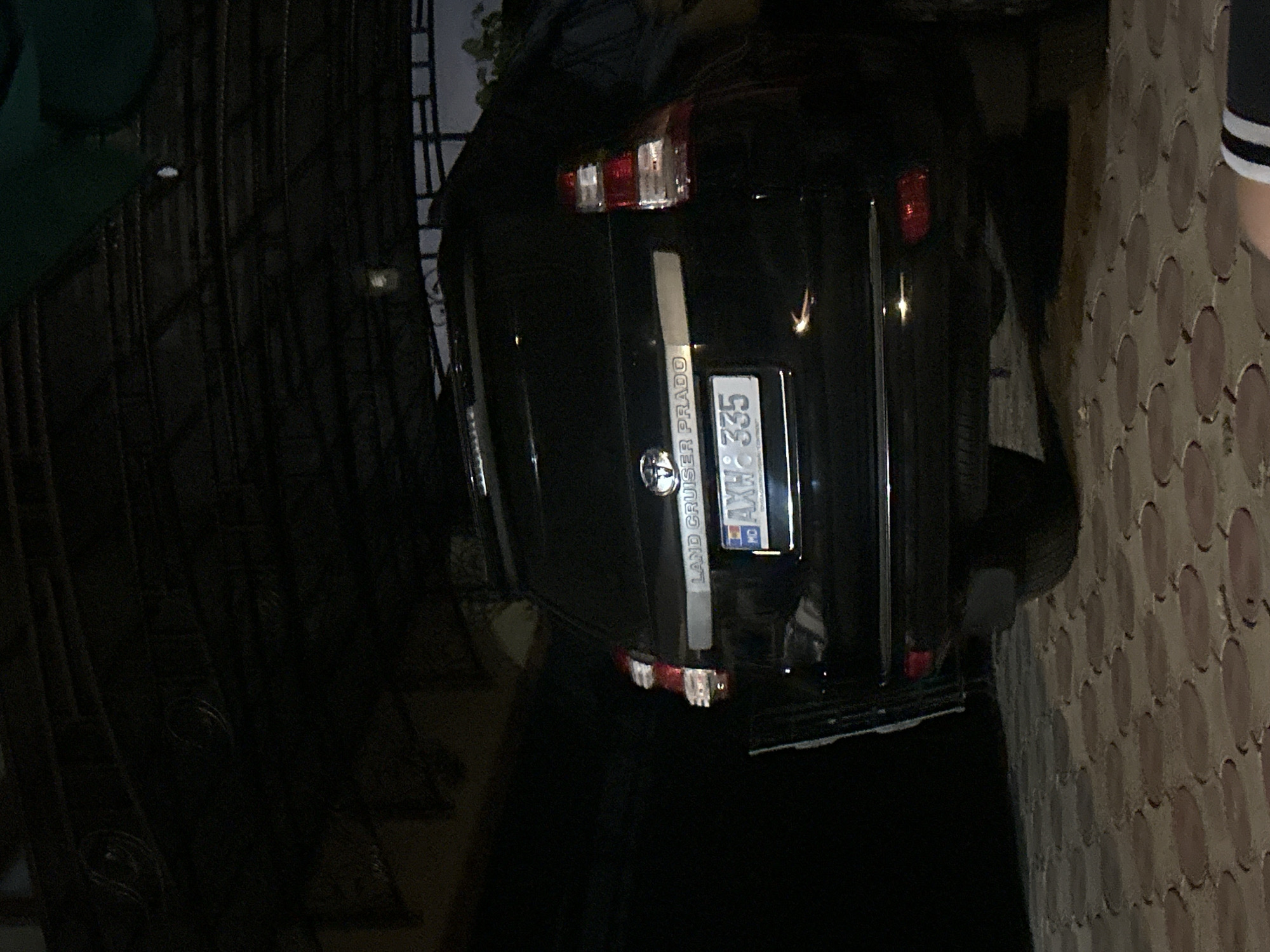 The image shows the rear of a black Toyota Land Cruiser Prado parked in a driveway or under a carport. The license plate is visible and reads "AXH 335". The car has a chrome trim piece above the license plate and the "LAND CRUISER PRADO" badge is also visible. The ground is paved with interlocking, circular brick pavers. The scene appears to be dimly lit, suggesting it is nighttime or an indoor shaded area. A dark green fabric is partially visible at the top of the frame. There are no people clearly visible in the image, though a hand with black and white stripes on it is visible in the lower right corner. The image has a slight tilt.