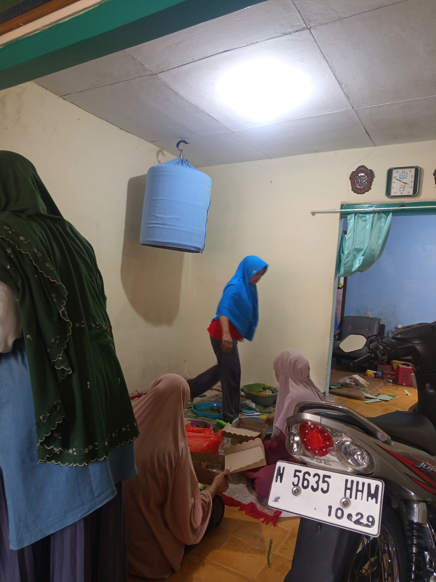 The image depicts an indoor scene, likely a residence, with several individuals present. The room appears somewhat cluttered. In the foreground, a portion of a motorcycle with a license plate reading "N 5635 HHM 10.29" is visible. Several people are seated or standing on the floor, most of whom are wearing hijabs or headscarves. One person in a blue hijab and red shirt is standing and appears to be moving across the room. Another individual in a green hijab and blue shirt is partially visible on the left side of the frame. A blue cylindrical object, possibly a bird cage cover, hangs from the ceiling. Various items are scattered on the floor, including what appear to be food containers, plastic bags, and some green leaves. In the background, a doorway with a light blue curtain leads to another room. Two clocks are mounted on the wall, and a decorative oval frame is also visible. The overall lighting suggests an interior setting with artificial light sources. The scene is consistent with a residential interior in Mulyorejo, Indonesia.
