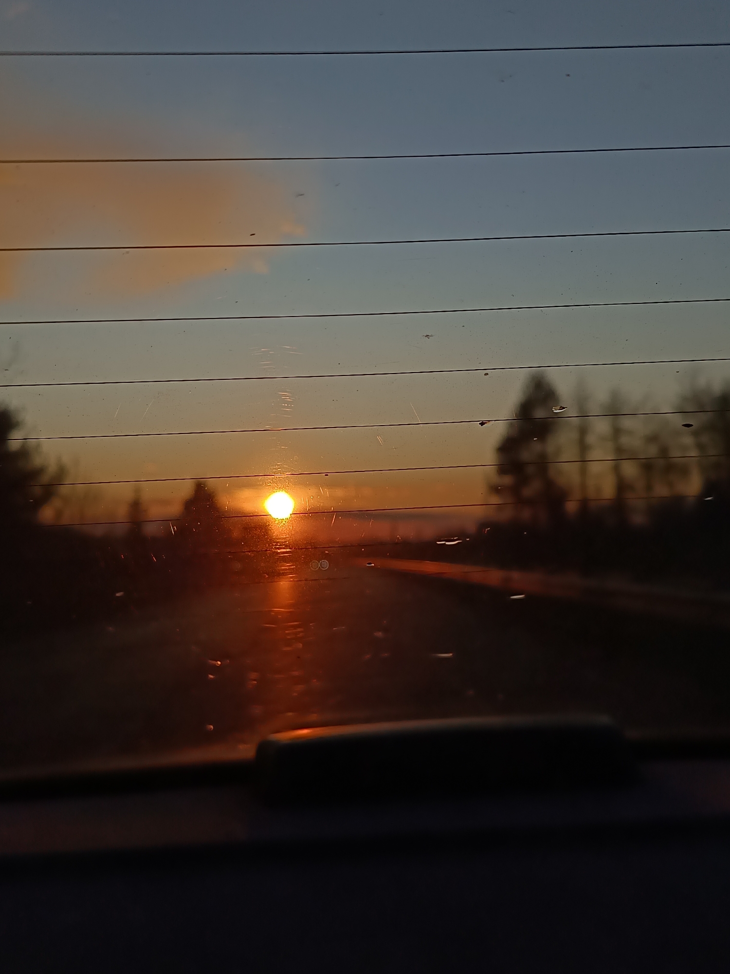 The image depicts a sunset observed through the rear window of a vehicle. The sun is situated near the horizon, emitting a bright, golden light that reflects onto the road surface in the distance. The window is lined with defroster filaments. Silhouetted trees and landscape elements are visible against the sky. The sky displays a gradient of colors, ranging from a light blue at the top to a deeper orange and yellow closer to the horizon. Part of the vehicle's interior is visible in the foreground. The location is indeterminate from the available information.