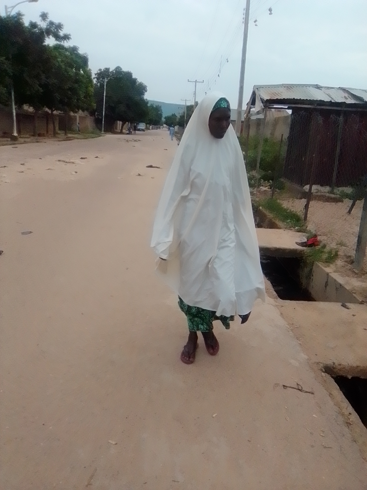 A full-body, eye-level shot shows a woman walking on a dirt road in Gombe, Nigeria, from a low-angle perspective. The woman is wearing a white hijab and a patterned green skirt, with dark sandals on her feet. She is walking away from the camera, towards the right side of the frame. Her arms are not visible, and her body is angled away from the camera. The road is unpaved and appears to be dusty, with some scattered debris. To the right of the road, there is a concrete drainage channel. In the background, there are trees, buildings, and utility poles. The sky is overcast and appears to be a light gray. The overall scene depicts a simple, everyday moment in an urban or semi-urban environment in Nigeria.