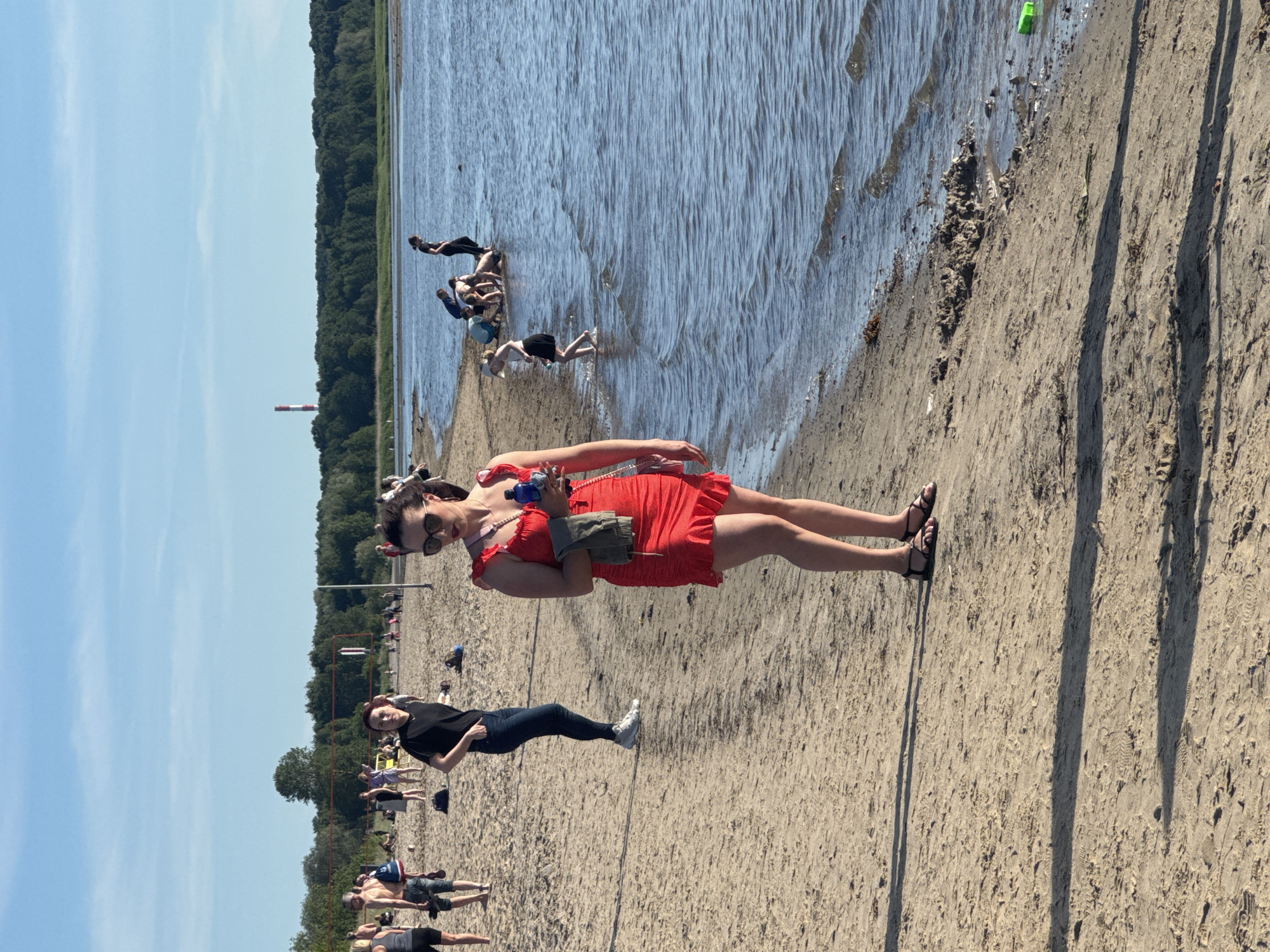 The image depicts a sunny day at a beach in Tallinn, Estonia. The foreground features a sandy beach with the edge of the water on the right. A woman in a bright red dress and sunglasses stands on the sand, holding a bottle of water and a folded cloth. In the background, other people are scattered on the beach and in the shallow water. Some individuals are playing volleyball, while others are wading or sitting near the water's edge. The water extends to a treeline in the distance, with a tall, red and white striped industrial chimney visible above the trees. The sky is clear blue with some wispy clouds.