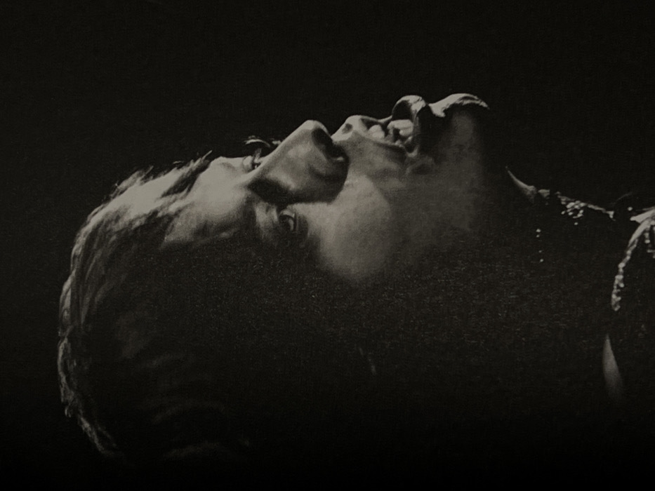 A monochromatic, close-up, profile portrait depicts a male individual with dark, layered hair, captured from the chest upwards, facing right. His right eye is partially visible, looking forward. His mouth is open wide, with the lower lip distinctly extended and a portion of his tongue and teeth visible, indicating an act of singing or vocalization. Dramatic lighting from the right illuminates the contours of his face, particularly his nose, cheekbone, and the right side of his mouth, while the left side of his face remains in deep shadow. A textured detail, possibly a beaded necklace or part of a garment, is faintly visible around his lower neck and upper chest area. The background is uniformly dark and featureless, providing no environmental context or specific location within Tel-Aviv, Israel, or any other city.