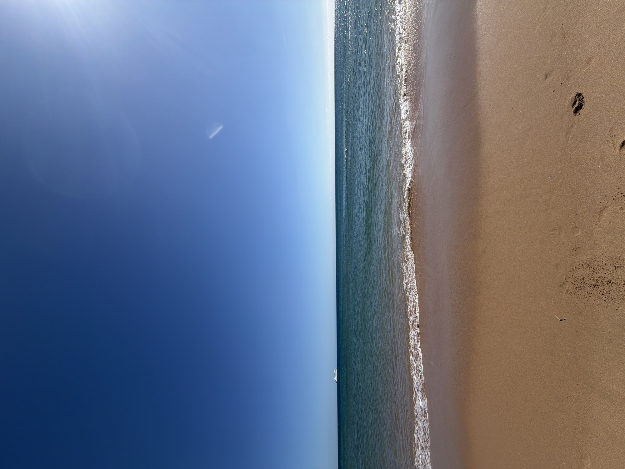 The image shows a beach scene, possibly Carcavelos Beach in Cascais, Portugal, characterized by a blue sky, calm water, and light-colored sand. The sky transitions from a dark blue to a lighter blue hue near the horizon. The water has gentle waves, marked by white foam. The sand appears clean with some scattered debris and footprints. A single white sailboat is visible on the horizon. There are no visible people in the image.