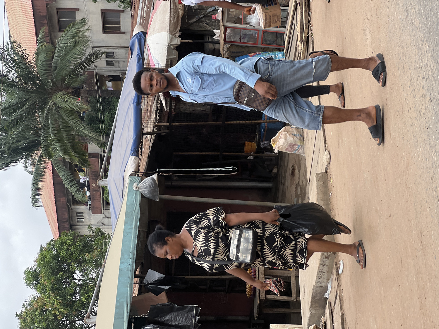 The image captures a street scene in Nkpor, Nigeria, featuring two pedestrians. The perspective is slightly elevated, looking down at the street and the people.
In the foreground, a man stands facing the camera. He wears a light blue long-sleeved button-up shirt, faded denim shorts, and black slides. A brown crossbody bag with a distinctive monogram pattern, resembling Louis Vuitton, hangs across his chest. He has short, dark hair and a neutral expression.
Behind and to the right of the man, a woman is seen walking away from the camera. She wears a black and white patterned dress, black flip-flops, and carries a black plastic bag in her left hand. A black shoulder bag, also featuring a monogram pattern similar to Louis Vuitton, hangs on her right shoulder. Her head is slightly turned to her right, and her expression is not clearly visible.
The setting is an outdoor street or market area. To the left, a large palm tree dominates the midground, with additional green foliage visible. Buildings with corrugated iron roofs and red roof tiles can be seen in the background, suggesting residential or commercial structures. Along the street, makeshift stalls or shops with blue and light blue/beige awnings line the path. Various items, including a white sack and a black plastic bag, are visible near the stalls. The ground is an unpaved or roughly paved surface.
The time of day appears to be daytime, likely overcast, as there are no harsh shadows. No distinct readable text is visible on signs or objects, apart from the monogram patterns on the bags. The scene depicts typical daily activity in an urban or semi-urban Nigerian environment.