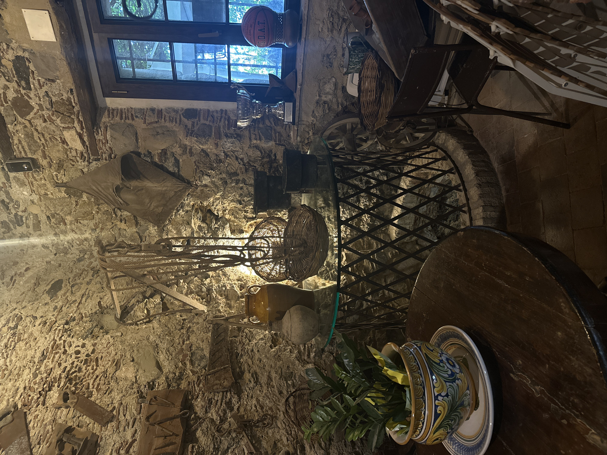 The photograph captures an interior scene in Rina, Italy, displaying a room decorated with an assortment of rustic objects. The walls are constructed of rough stone. A window is at the top of the image. A variety of items, including baskets, a glass-topped table with a metal base, and a wooden table with a plate and an ornate vase filled with greenery, are displayed. Agricultural tools and old wooden objects are hung against the stone walls. A basketball is present in the background near the window.