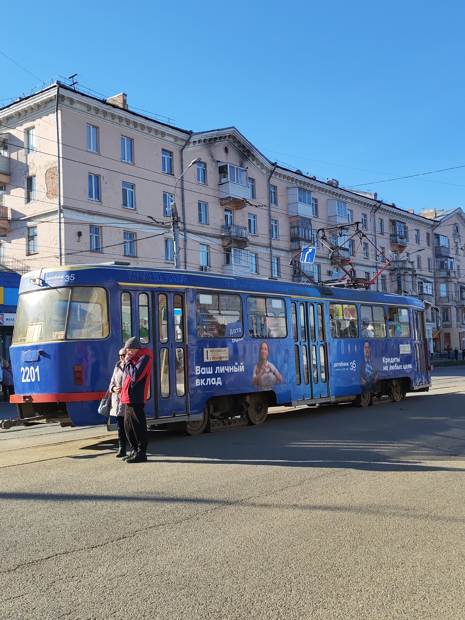 A blue tram, numbered 2201 and displaying route 35, is stationary on a city street in Izhevsk, Russia, under a clear blue sky. The tram is adorned with advertisements for "Databank," featuring Russian text such as "Ваш личный Вклад" (Your personal deposit), "Кредиты на любые цели" (Loans for any purpose), and "ИЗУЧИТЕ ВСЕ УСЛОВИЯ" (Study all conditions). The ads also mention "Без кондуктора" (Without conductor) and "МОСКОВСКАЯ Г. МЕТАЛЛУРГОВ" (Moscow, G. Metallurgov), likely an address or branch.
Two people are standing near the front left side of the tram. One individual wears a red jacket and a hat, while the other is in a light-colored jacket. Their shadows are cast long on the asphalt, indicating bright sunshine and possibly morning or late afternoon.
In the background, a light pink/peach colored multi-story building with numerous windows and balconies lines the street. Its architectural style suggests a mid-20th-century urban environment. Overhead lines for the tram and power lines crisscross the sky, and a blue road sign with a white arrow is visible above the tram. The scene depicts a typical, sunny day in an urban setting.