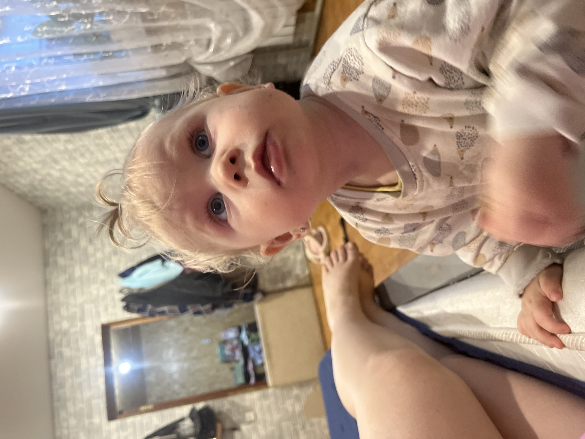 A toddler with blonde hair and blue eyes is looking directly at the camera. The child is wearing a light-colored long-sleeved pajama top with a pattern of stylized hedgehogs or similar creatures. Part of an adult's legs and feet are visible in the lower portion of the frame, suggesting the child is on a lap or near an adult.
In the background, there's a domestic interior. A mirror reflects a view of clothing hanging in what appears to be a bedroom or living area. A beige-colored storage cube is also visible. Parts of a bed or sofa are evident.
The image appears to have been taken indoors, possibly in a private home. It is not possible to ascertain the specific location as Vesyoloye, Russia, from the image alone.