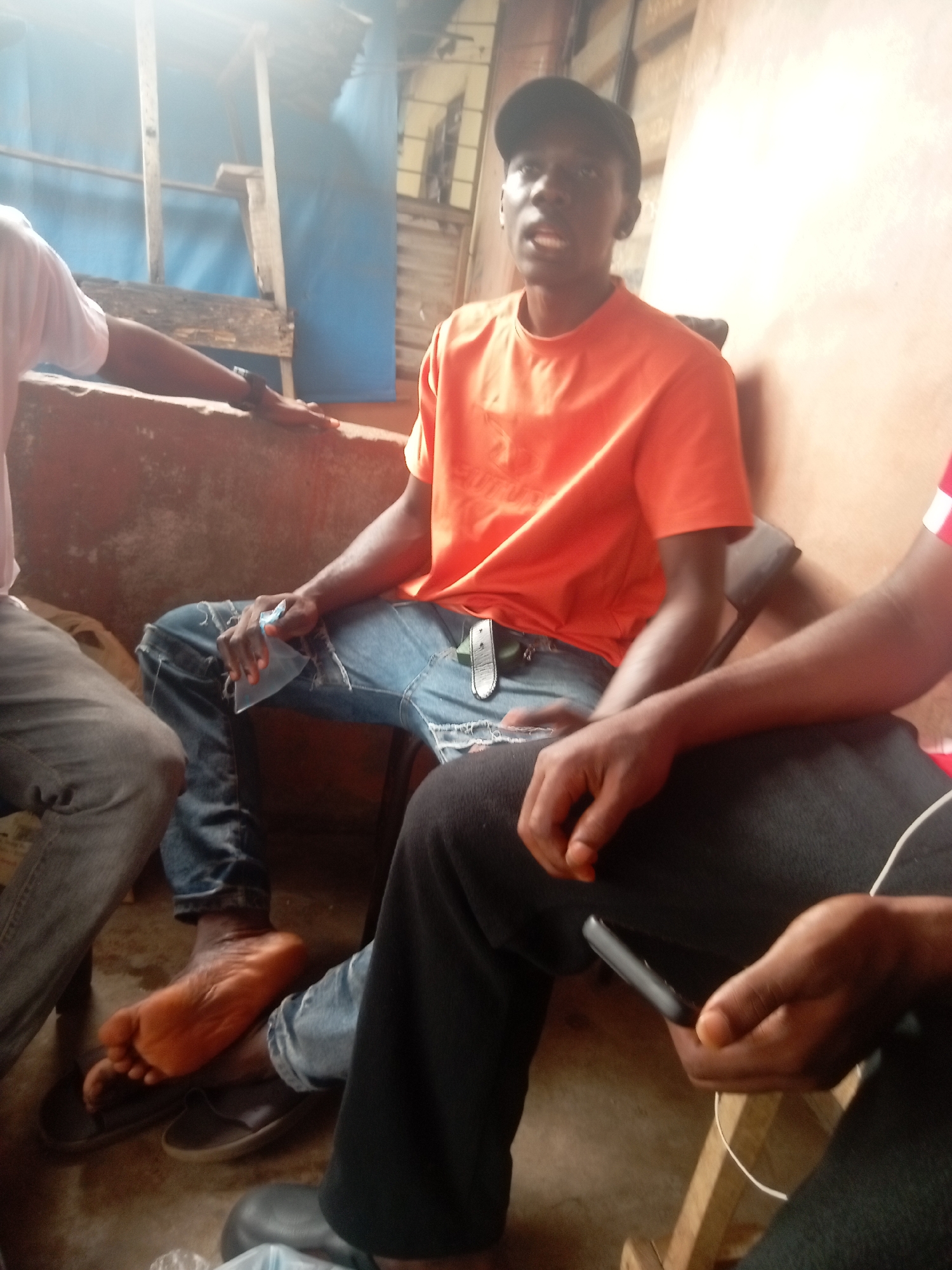 The image depicts four individuals in an indoor setting in Nkpor, Nigeria. The central figure, a young Black man, is seated on a chair, wearing an orange t-shirt and ripped jeans with a black cap. He appears to be speaking. To his left, partially visible, is an individual wearing a white shirt and gray jeans, with one foot bare and resting on a sandal. To the right of the central figure is another individual wearing black pants. This person is holding a black smartphone in their hand. The background includes a concrete wall. The presence of exposed wooden beams, and blue tarpaulin suggests a semi-constructed or unfinished building. The bright natural light further indicates an outdoor or semi-outdoor environment.