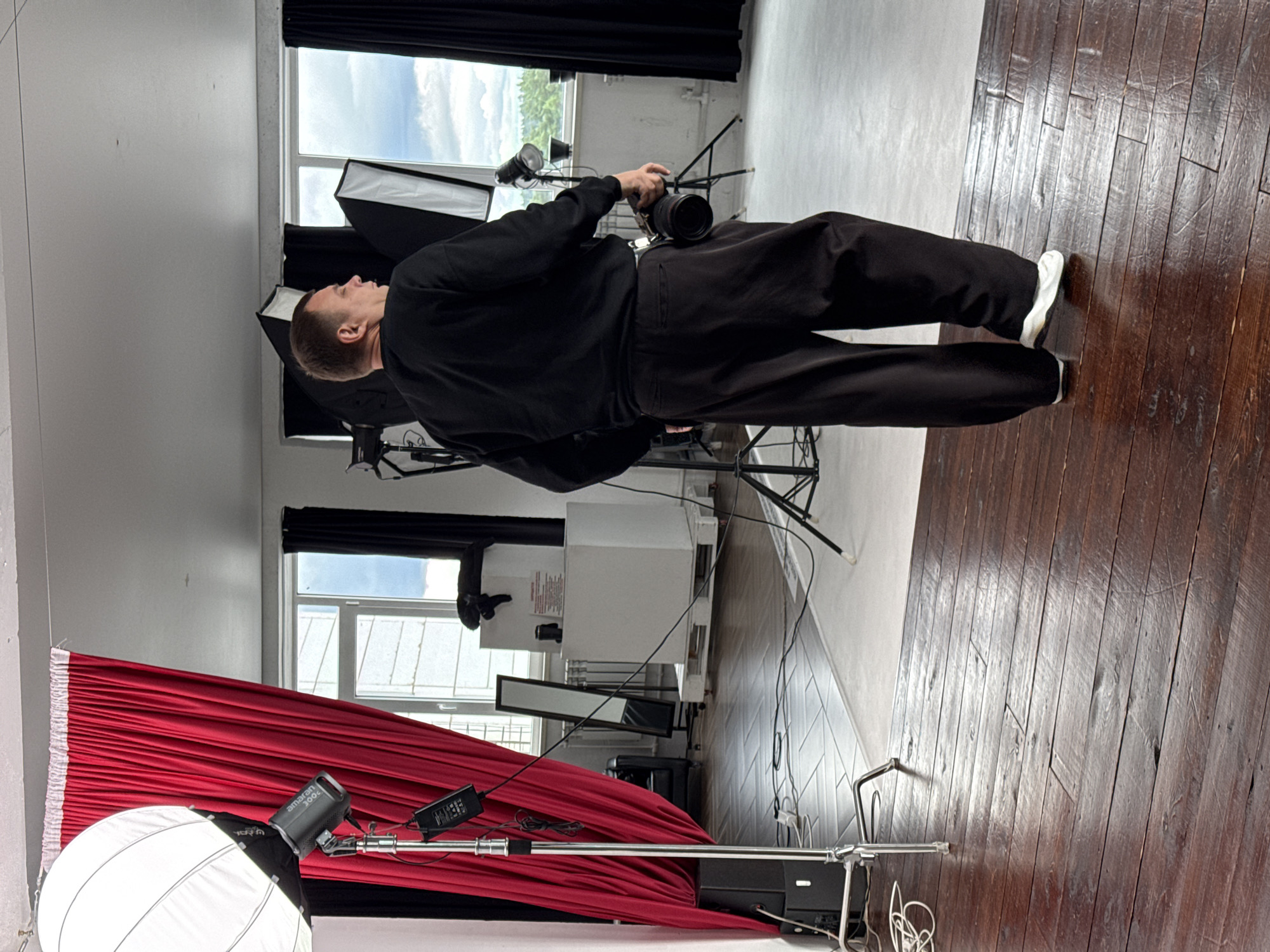 A person stands in a photography studio, facing away from the camera. They are wearing a black sweatshirt and dark, wide-legged pants, and white sneakers. A camera with a lens is attached to their hip. The studio has a dark wooden floor and a white floor in the background. To the left, a large white softbox is mounted on a stand, partially obscured by red curtains. Various studio lights and equipment are visible throughout the space. Large windows with a view of greenery and a cloudy sky are present in the background. The studio appears to be located in Chaikovsky, Russia, as indicated by the provided information.